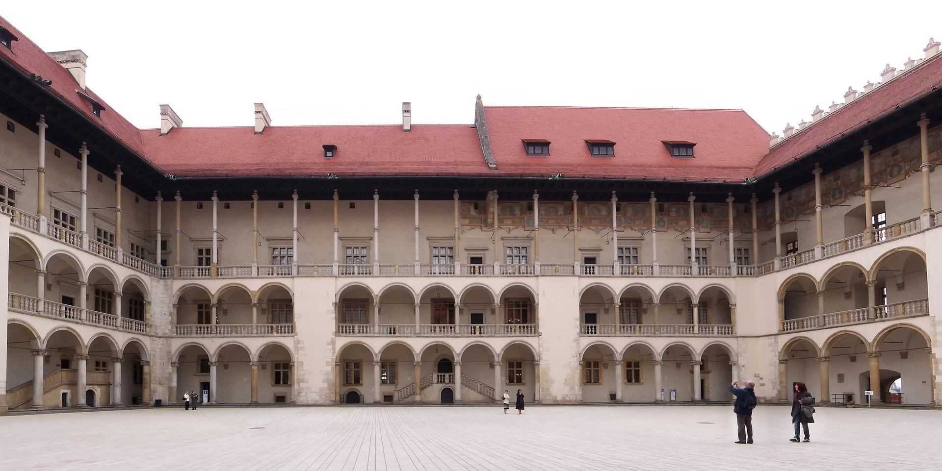Kliknij, aby powiększyć Zdjęcie wewnętrznego dziedzińca zamku. Wawel otoczony jest z trzech stron zabudowaniami. Budynek ma trzy kondygnacje. Na parterze i pierwszym piętrze widoczne są krużganki. Na ostatnim piętrze są liczne filary podtrzymujące dach. Otwory okienne są przedzielone szprosami na kilka mniejszych okien. Dach jest dwuspadowy.