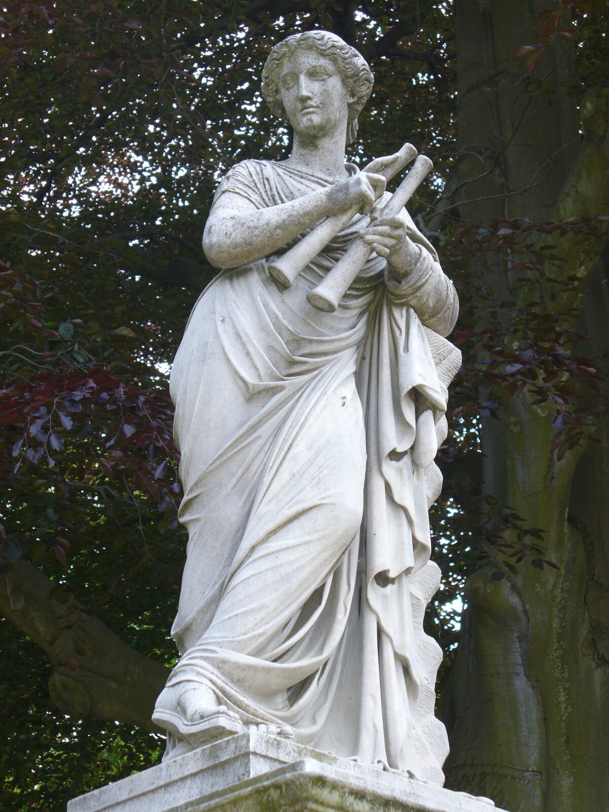  Kolorowa fotografia przedstawia marmurową rzeźbę młodej kobiety o falowanych, spiętych włosach. Ubrana jest w grecką tunikę, która okrywa jej ciało od szyi, aż do kostek stóp. Na nogach ma sandały. Trzyma w dłoniach dwa długie instrumenty w kształcie piszczałek, które mają cylindryczny kształt i długość ok 30‑40 cm. Rzeźba ustawiona jest na podwyższeniu, w tle pień i gałęzie pokryte liśćmi rozłożystego drzewa.  