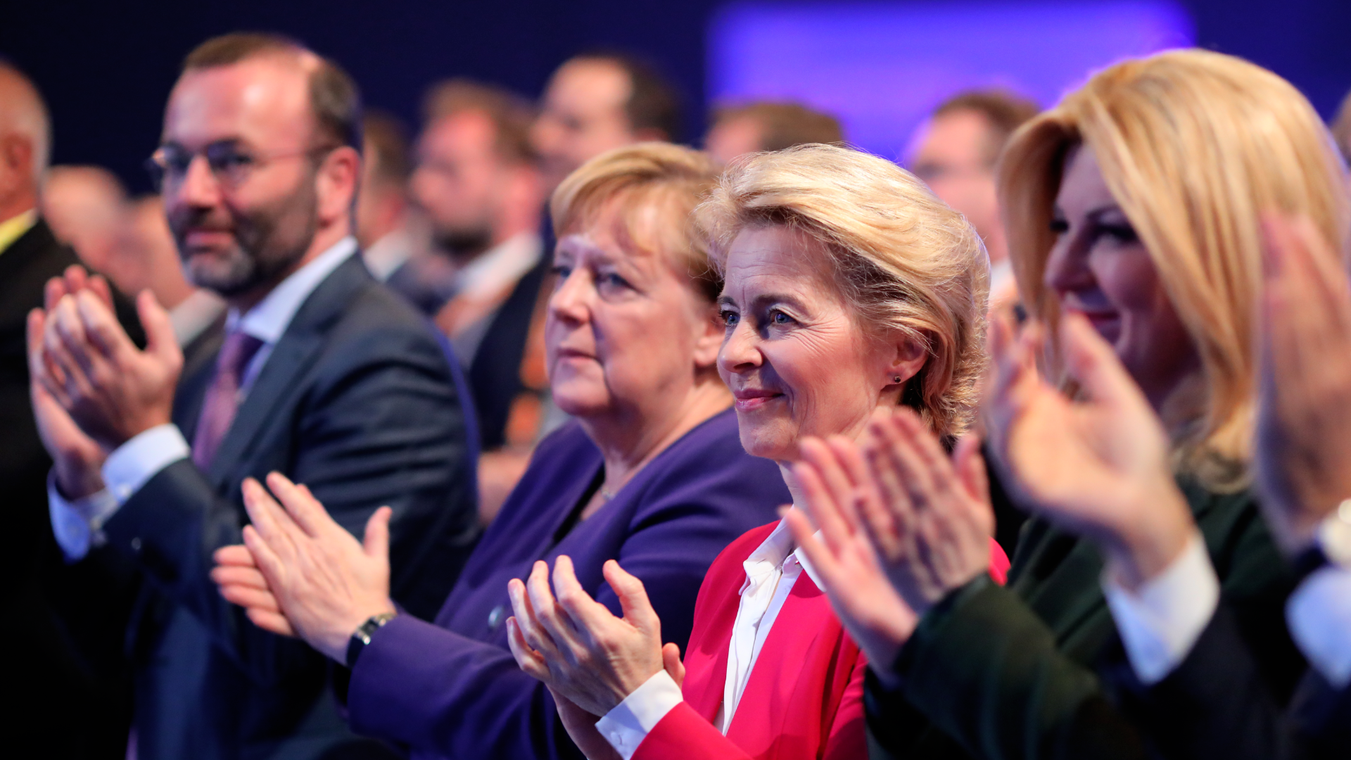 Zdjęcie przedstawia grupę ludzi, którzy biją brawo. Ich wzrok skierowany jest w jedną stronę. Na pierwszym planie znajdują się trzy kobiety. Wśród nich: Angela Merkel i Ursula von der Leyen.