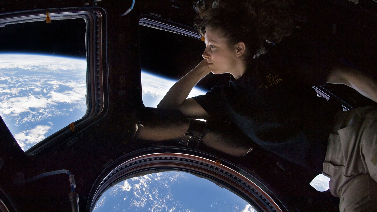 Zdjęcie przedstawia młodą kobietę w statku kosmicznym, Po prawej stronie widoczna jest leżąca na prawym boku kobieta, ubrana w czarną bluzkę z krótkim rękawem i brązowe spodnie. Kobieta prawym łokciem opiera się framugę okna, a dłonią podpiera głowę. Spogląda w lewo  i podziwia widok na kulę ziemską, który roztacza się z okien statku kosmicznego.