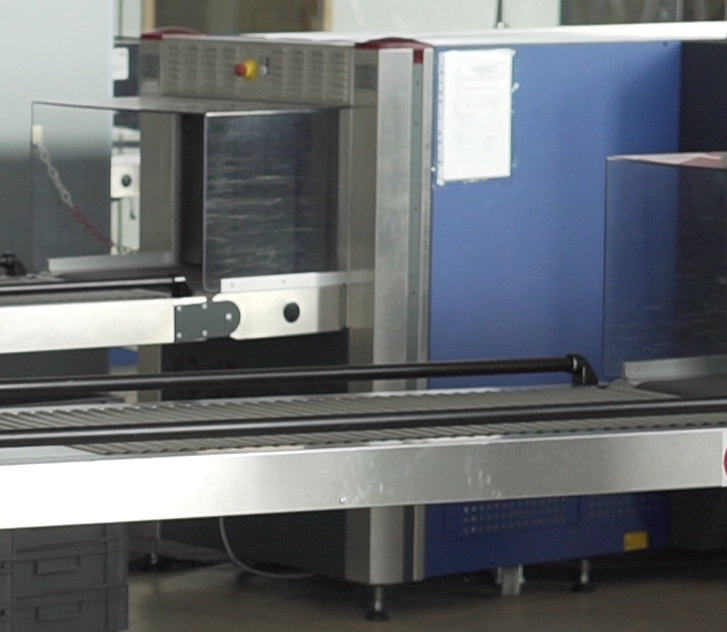 Zdjęcie przedstawia urządzenie do prześwietlania bagażu. Widoczna jest skrzynia urządzenia, do której otworu, osłoniętego przezroczystymi płytami, prowadzi taśmociąg. Otwór zasłonięty jest gumowymi pasami zwisającymi z jego szczytu. Nad otworem znajduje się czerwony przycisk awaryjnego zatrzymania urządzenia.