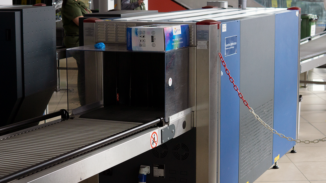 Zdjęcie przedstawia iks rej er te gie, czyli urządzenie do prześwietlania bagażu. Jest to duże i długie urządzenie, które stoi na podłodze. Z jego dwóch stron wychodzi taśmociąg. Urządzenie przypomina wielkie, szaro niebieskie pudło, do którego za pomocą taśmociągu wchodzi i wychodzi bagaż. Z boku siedzi pracownik i na ekranie przed sobą ma pokazany prześwietlony bagaż.