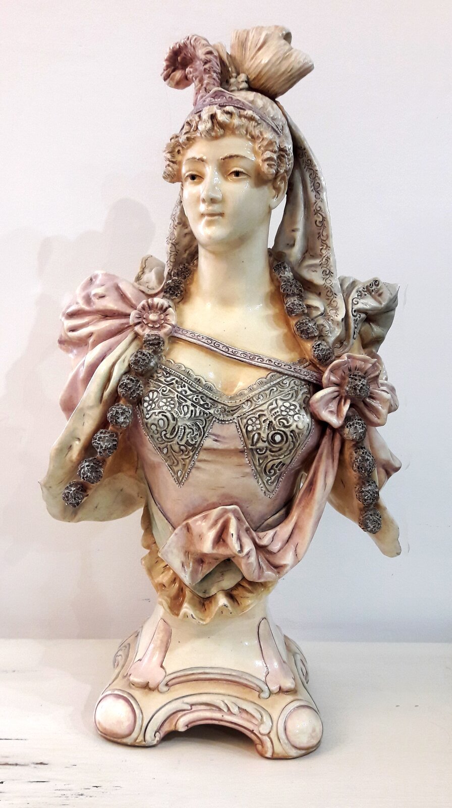 Ilustracja przedstawia popiersie kobiety, ubranej w bogaty strój w różowych odcieniach, dekorowany koronkami. Jej głowa jest lekko uniesiona i dumna. Zdobi ją pióropusz.