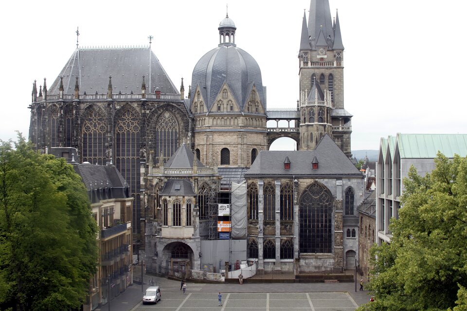 Fotografia przedstawia widok na bryłę Katedry  w Akwizgranie. Budynek kaplicy składa się z dwóch kondygnacji oraz wysokiego dachu, na którym znajduje się dzwonnica. Kaplica zbudowana jest z jasnego kamienia, zaś sam dach to szara dachówka, która wpasowuje się do koloru budynku. Obok kaplicy widoczny jest plac i drzewa oraz odpoczywający ludzie. Fotografia została wykonana w lekko pochmurny dzień. 