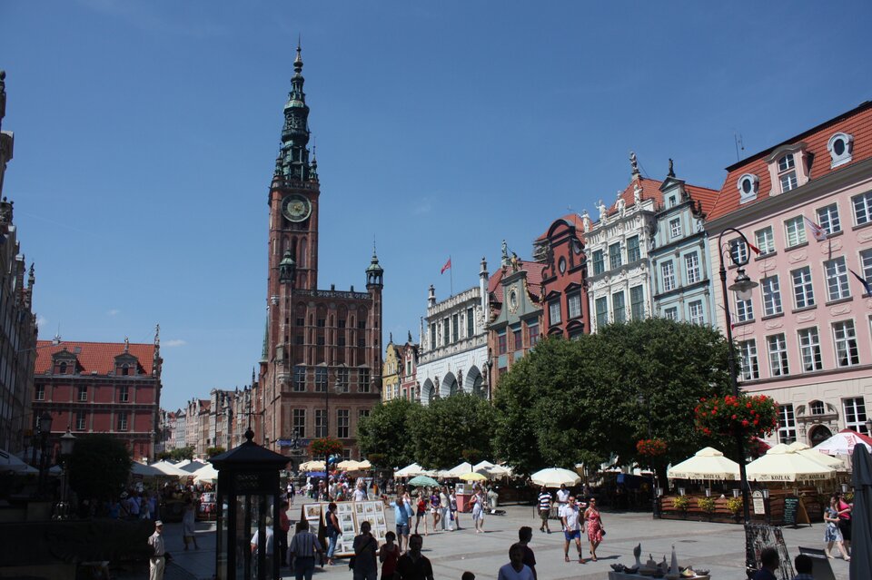 Na zdjęciu widoczna jest starówka Gdańska ze starymi zabytkowymi kamienicami i ustawionym w centralnej części kadru szesnastowiecznym ratuszem z wysoką wieżą zegarową. Ratusz zbudowany jest w stylu niderlandzkim manierystycznym. Ma kolor rudo‑jasnobrązowy, zbudowany jest z kamienia i cegły. Kopuła wieży jest jasnoturkusowa. Od prawej strony kadru przez całe zdjęcie ciągną się usytuowane jedna obok drugiej wysokie, zadbane kamienice w różnych kolorach. Niektóre z nich pokryte ornamentami i zdobieniami. Przed kamienicami, na deptaku, po którym spacerują osoby ubrane w letnie stroje, ustawione są kawiarniane parasole. Część z nich zasłaniają drzewa. Po lewej stronie kadru także są kamienice i parasole, nie są one jednak tak widoczne. Ta część kadru jest zacieniona. W tle bezchmurne niebo.