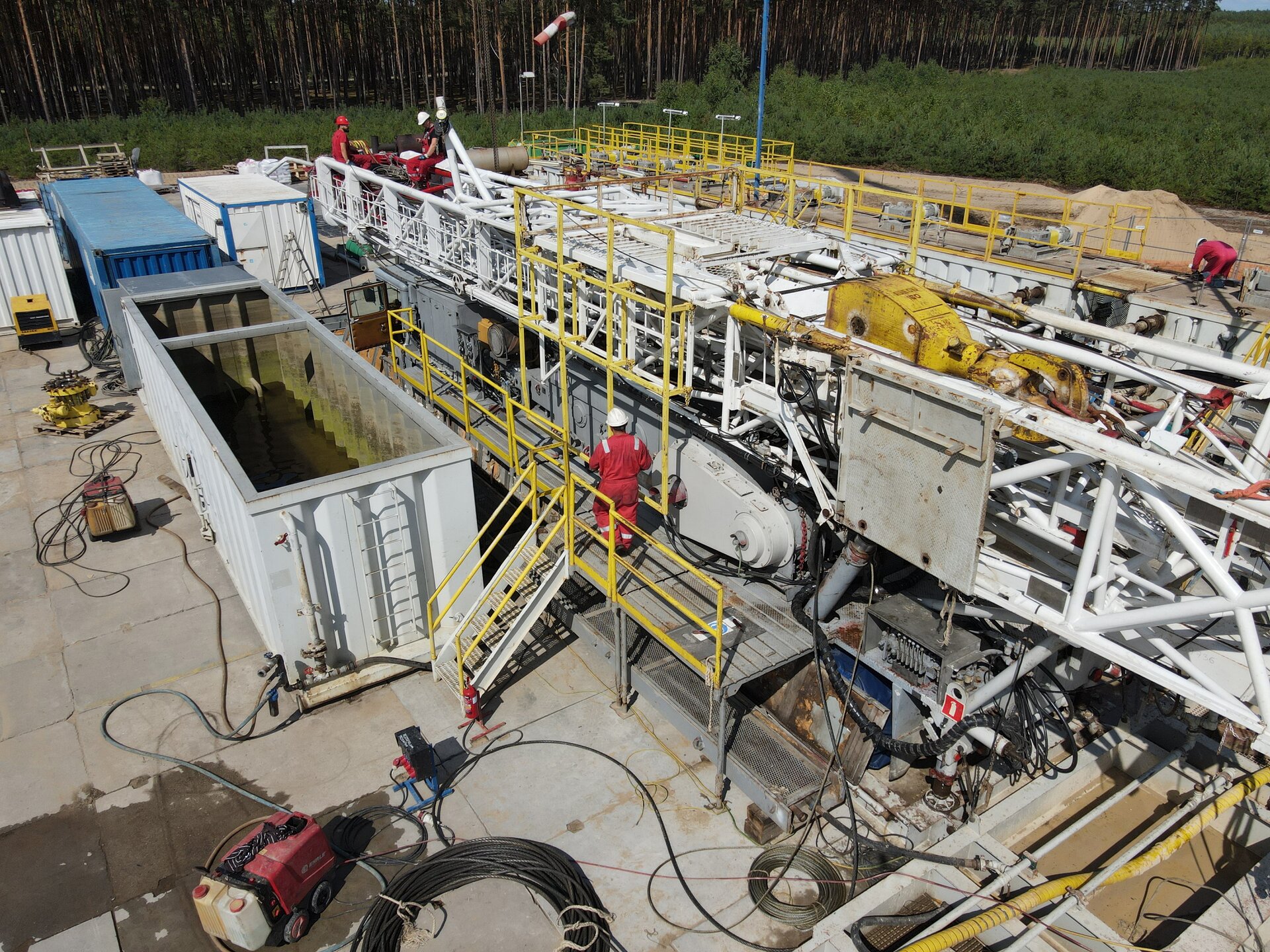 Na zdjęciu widoczny jest maszt wiertniczy w trakcie przygotowywania do podnoszenia. Centralnym elementem zdjęcia jest betonowy plac wiertniczy. Na placu rozłożony jest podest. Na podeście widoczni są dwaj robotnicy ubrani w czerwone stroje ochronne i białe kaski. Na podeście leżą fragmenty stalowej konstrukcji kratowej. Jest to maszt w trakcie przygotowania do podniesienia. Po lewej stronie znajduje się biały prostopadłościenny zbiornik przypominający kontener.W tle zdjęcia widać drzewa.