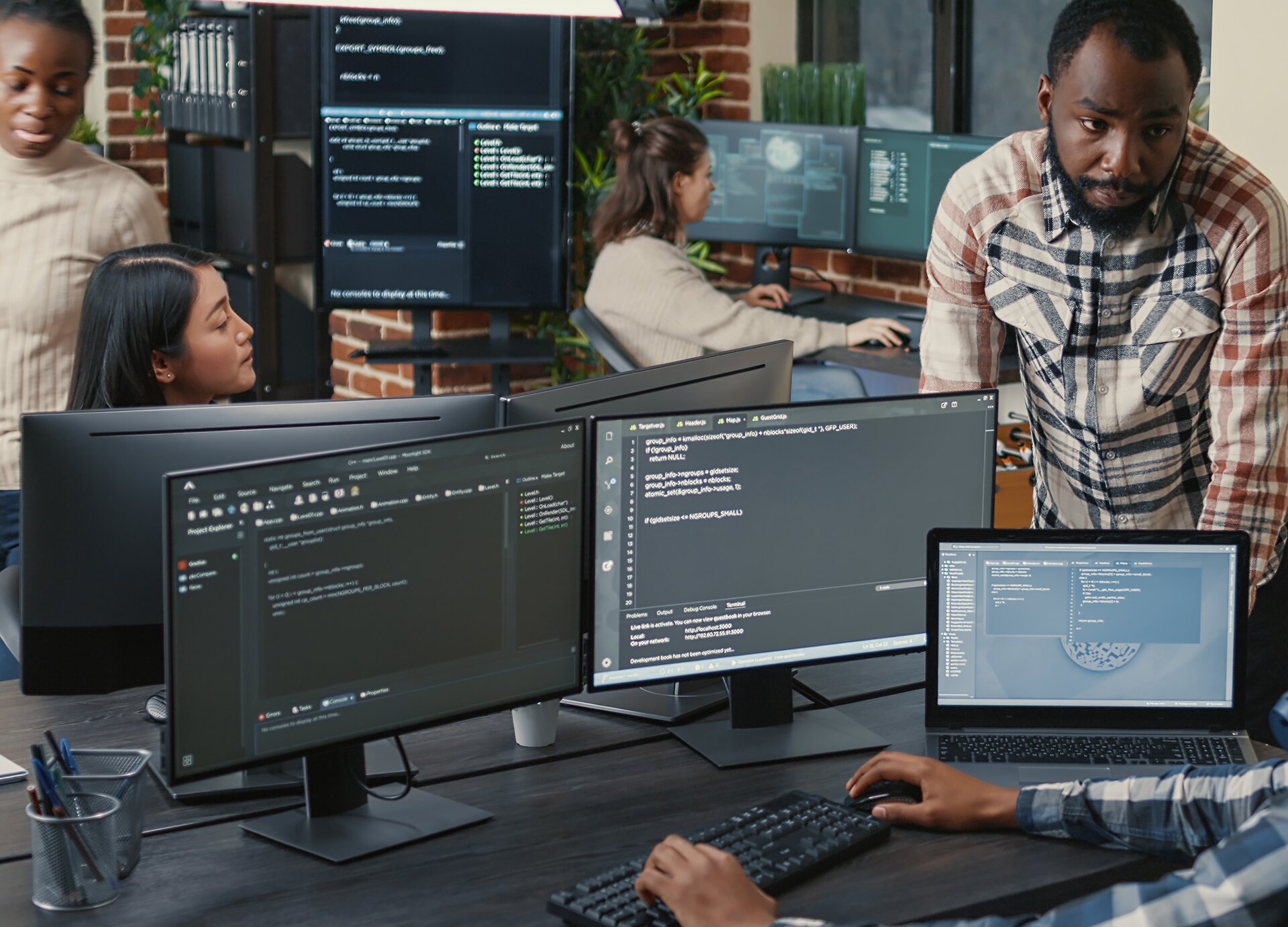 Zdjęcie przedstawia wnętrze pomieszczenia biurowego, w którym widoczne są biurka, na których stoją duże monitory komputerów stacjonarnych oraz laptop. Na pierwszym planie widoczne jest biurko z dwoma monitorami oraz laptopem. Na ekranach widoczne są programy do programowania. Po prawej stronie widoczny jest fragment osoby siedzącej przy biurku i trzymającej obie dłonie na klawiaturze. Na prawo od biurka stoi mężczyzna, który opiera się rękami o blat i patrzy w stronę osoby siedzącej. Na drugim planie widoczna jest głowa kobiety patrzącej w prawo, wystająca zza dwóch monitorów komputerów. Za nią widoczna jest stojąca kobieta. W tle widać kolejne biurko z monitorami i siedzącą przy nim kobietę.