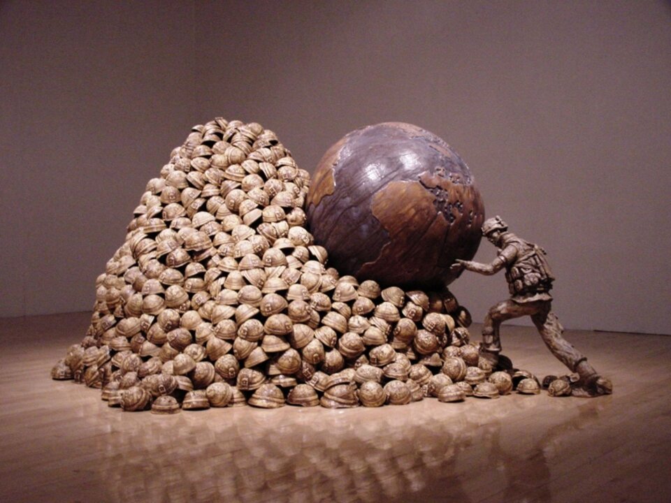 Zdjęcie przedstawia ceramiczną rzeźbę. Ukazuje ona żołnierza pchającego kamień w formie kuli ziemskiej pod górę zrobioną z hełmów wojskowych. Podpiera on kulę głową i rękoma.