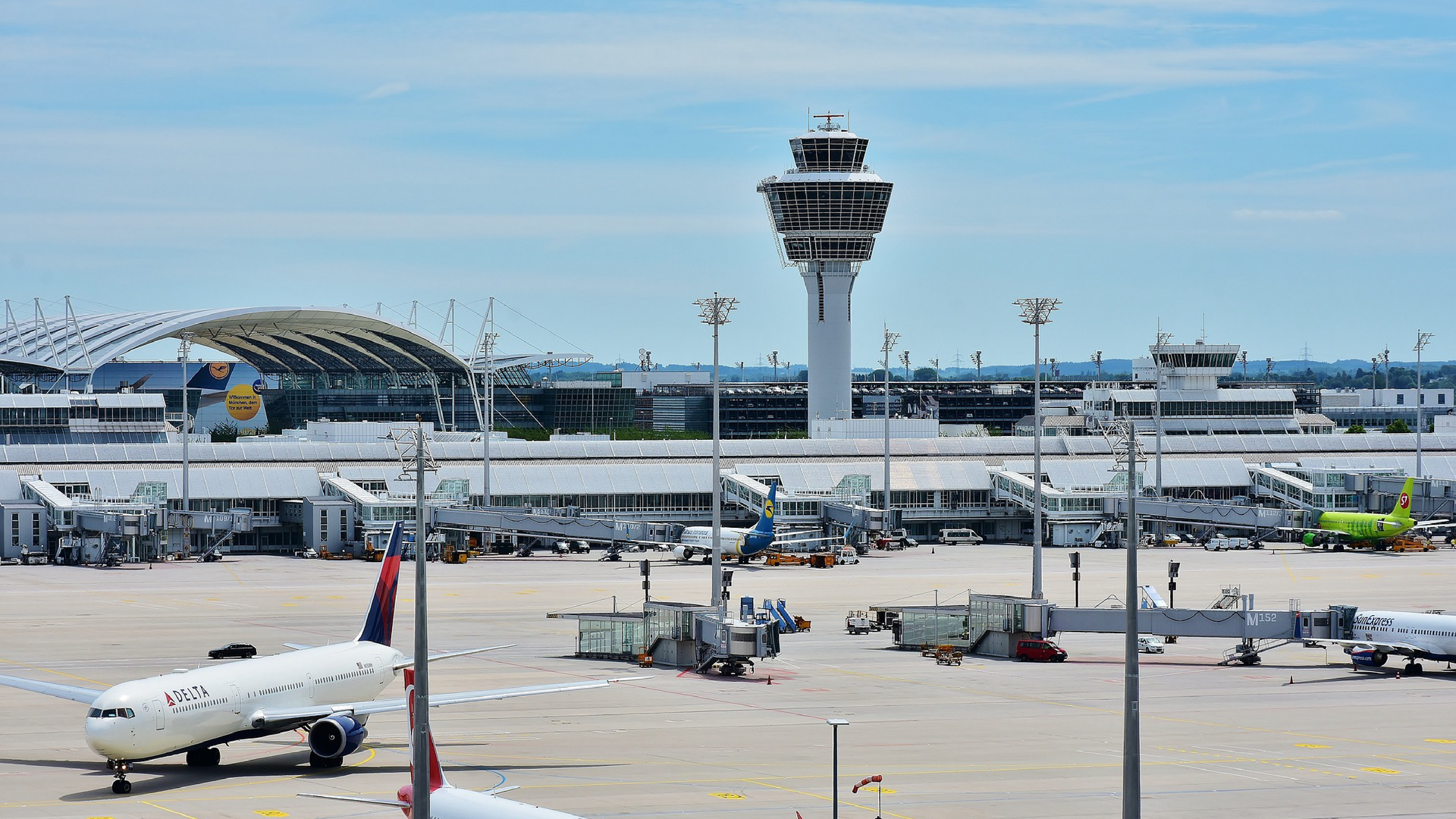 Zdjęcie przedstawia port lotniczy. Widoczne są samoloty podczas procedury wejścia pasażerów na pokład oraz samoloty stojące na płycie lotniska. Najwyższym obiektem na zdjęciu jest wieża kontroli lotów.