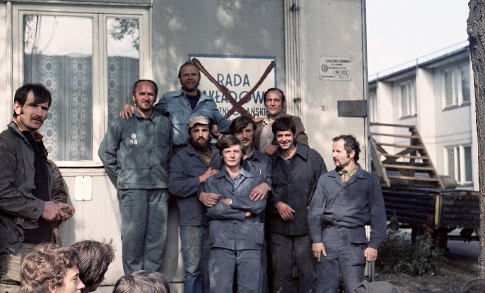 Zdjęcie przedstawia dwunastu pracowników Stoczni. Są to osoby strajkujące. W większości mają ubrane granatowe stroje robocze. Ośmioro spośród robotników zakrywa przekreśloną tablicę Rady Zakładowej, przymocowaną do ściany. W tle zdjęcia znajduje się przyczepa z drabiną oraz niskie zabudowania. W oknach budynków są firany.