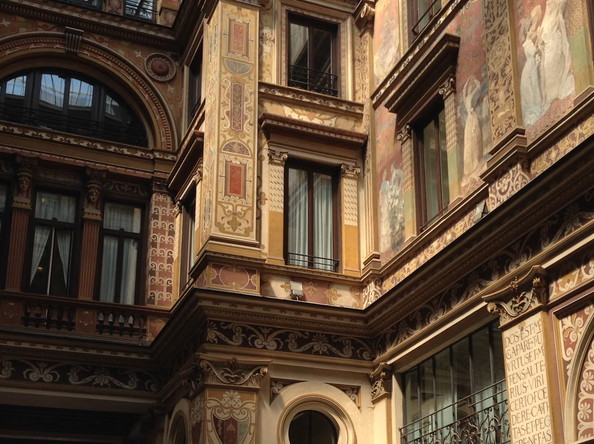 Grafika przedstawia malowidła ścienne na elewacji kamienicy: widoczne są postaci ludzkie, motywy florystyczne, napisy oraz inne elementy zdobnicze.