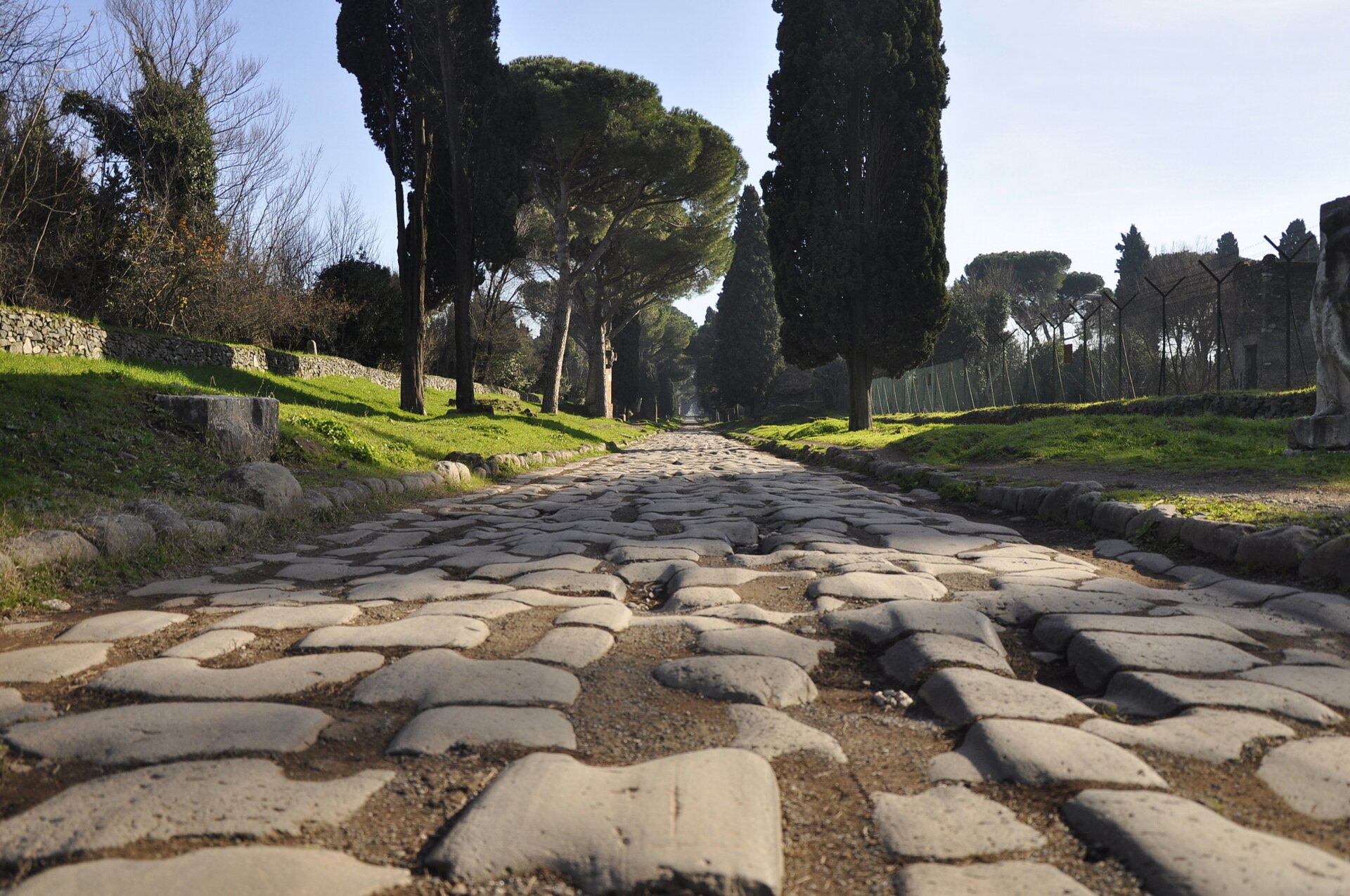 Ilustracja przedstawiająca rzymską drogę  Via Appia. Droga jest wyłożona kamieniami. Po obu jej stronach znajdują się drzewa i połacia trawy, zaś w oddali jest las.