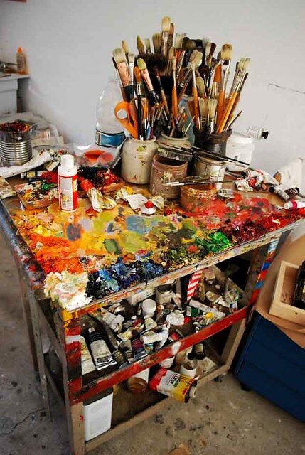 Na zdjęciu stoi niewielki stół z kwadratowym blatem, z dwiema półkami poniżej. W prawym górnym rogu stołu stoją słoiki z pędzlami różnej wielkości. Przed nimi trzy puste puszki, na których leżą pędzle. Na stole leżą niedbale położone tubki z farbą. Cały blat stołu pokryty jest różnej wielkości plamami po farbie. Na środku żółte, po bokach czerwone, niebieskie i zielone. Malarz musiał rozrabiać na nim farby lub wycierać pędzle. Na półkach pod blatem leżą w nieładzie tubki z farbami i pojemniki z olejem i terpentyną. W tle biała ściana, przy której stoją metalowe puszki, wiszą jakieś szmatki.