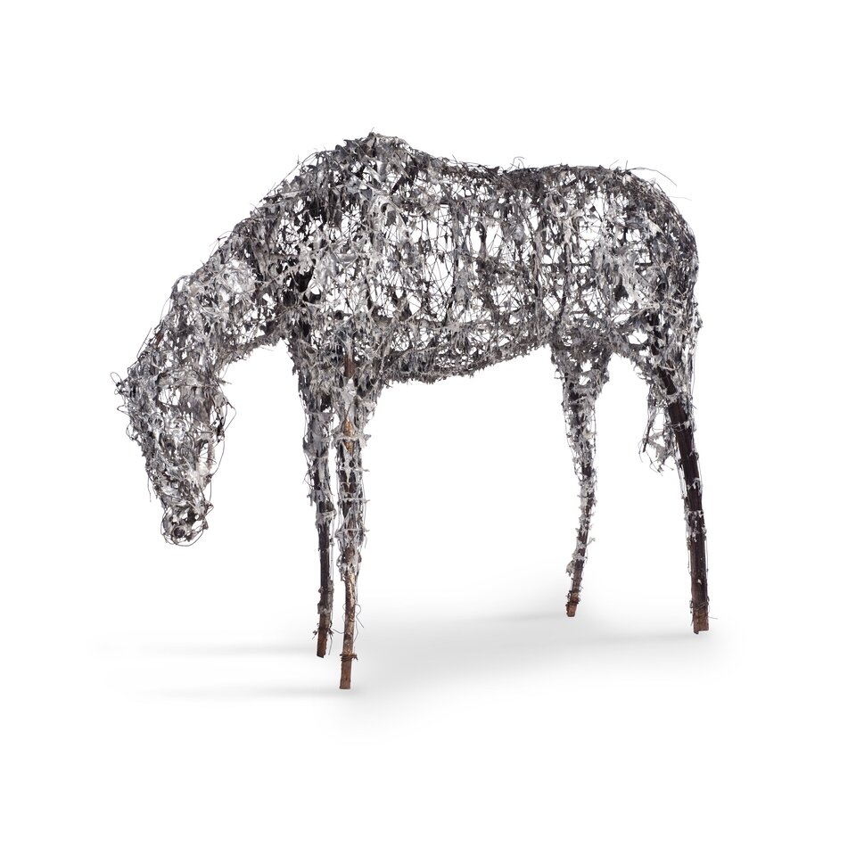 Fotografia przedstawia rzeźbę konia z pochyloną głową, pozbawionego kopyt oraz uszu. Korpus wykonano ze splecionego drutu oraz fragmentów metalu. Kończyny konia wykonano z tyczek oplecionych drutem. Tło, na którym zaprezentowano rzeźbę jest białe.
