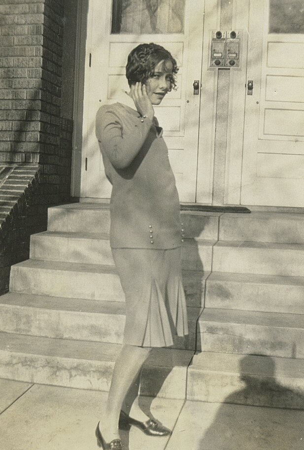 Zdjęcie przedstawia młodą kobietą, która stoi przed wejściem do budynku. Ma krótkie włosy, bluzkę z długim rękawem oraz spódnicę sięgającą do kolan. Kobieta patrzy w stronę widza i dotyka twarzy prawą ręką.