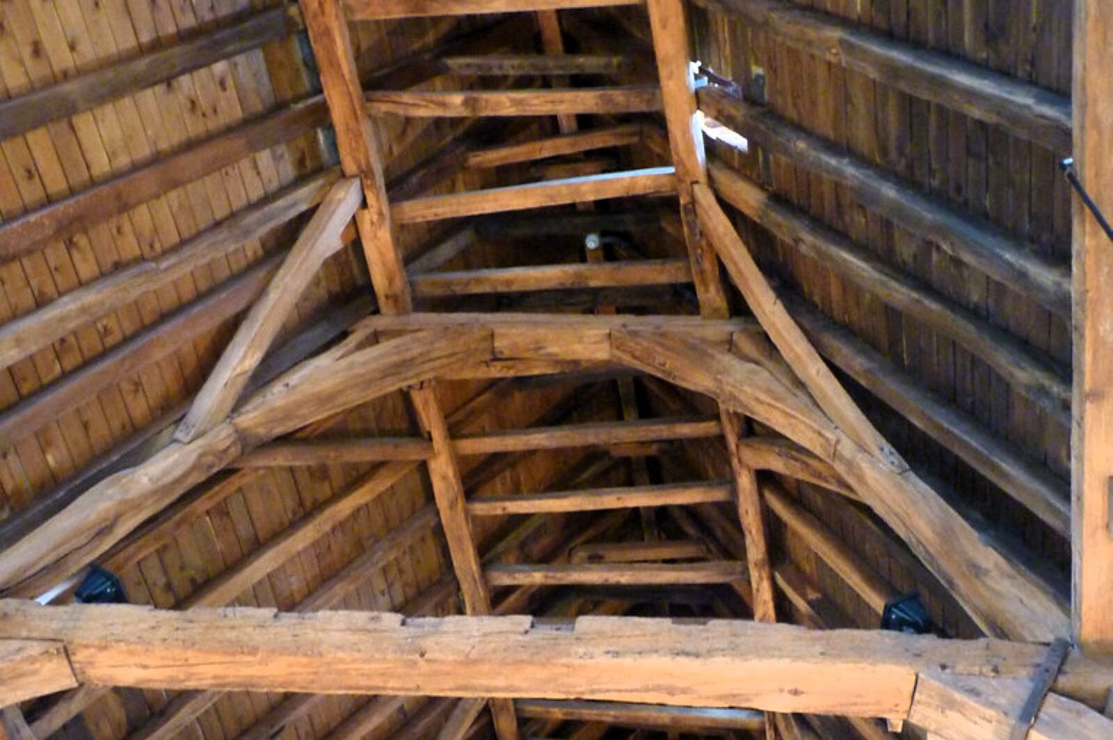 Zdjęcie przedstawia konstrukcję dachu z drewnianych belek. Jest to dach dwuspadowy, składający się z dwóch płaszczyzn (połaci dachowych) połączonych krawędzią na górze (tzw. kalenicą). Fotografia wykonana z dołu ukazuje konstrukcję z równolegle umieszczonych belek na płaszczyznach dachu i wzmocnieniu na samej górze, w formie przypominającej drabinę. Dodatkowo konstrukcja została wsparta grubszymi belkami połączonymi w formie dużej litery "A".