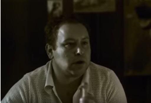 Zdjęcie przedstawia dojrzałego mężczyznę w jasnej, rozpiętej koszuli. Ma krótkie, ciemne włosy i twarz z drugim podbródkiem.