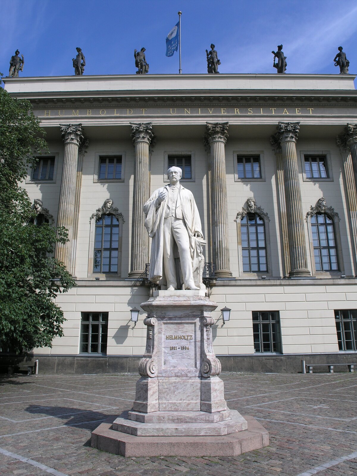 Na zdjęciu fragment klasycystycznego budynku uniwersyteckiego. Środkowe okna w budynku wykończone są od góry kamiennymi popiersiami. Pomiędzy oknami znajduje się rząd kolumn korynckich. Na środku dachu znajduje się maszt, na którym powiewa uniwersytecka flaga. Na dachu znajdują się również kamienne figury ludzkich postaci ustawione rzędem. Na pierwszym planie przed budynkiem ustawiony jest pomnik stojącego mężczyzny. Po lewej stronie rosną drzewa.