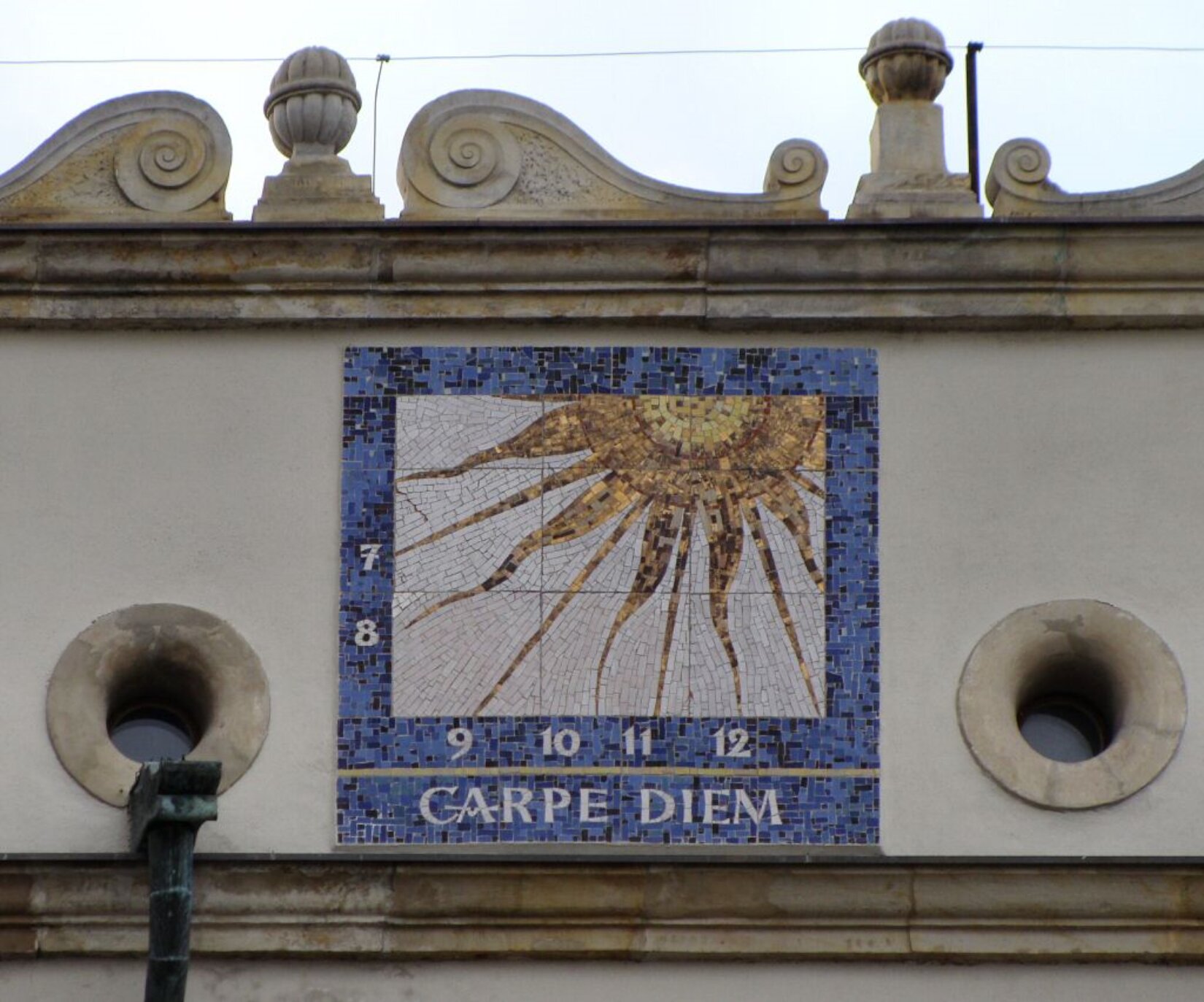 Fotografia przedstawia fragment elewacji Zamku Książąt Pomorskich, na której umieszczona została kolorowa mozaika prezentująca zegar słoneczny. Mozaika składa się z małych czworokątów w kolorach: niebieskim, czarnym, szarym, żółtym, brązowym oraz odcieniach wymienionych kolorów. Mozaika przedstawia niebieskie tło, wewnątrz którego znajduje się szare tło oraz słońce wraz z długimi promieniami. Na niebieskim tle umieszczone zostały cyfry: 7, 8, 9, 10, 11, 12. Poniżej napis: Carpe Diem.