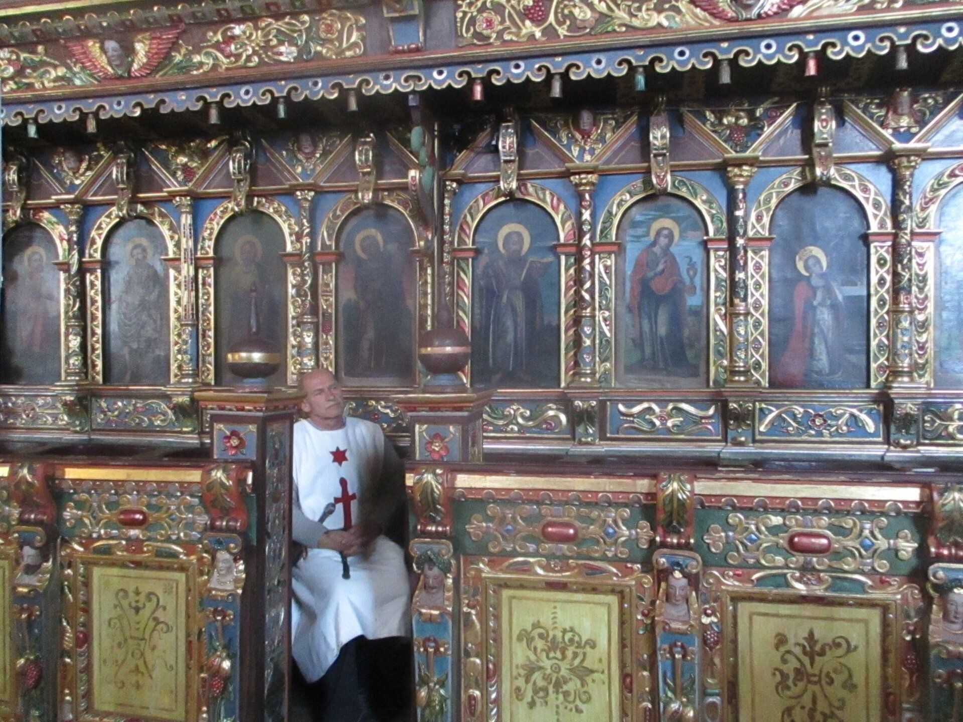 Zdjęcie przedstawia bogato zdobione prezbiterium. Cała ściana ozdobiona została obrazami przedstawiającymi sylwetki świętych. Pomiędzy obrazami, a także nad nimi, umieszczone zostały złoto‑czerwono‑niebieskie zdobienia. Pod obrazami znajdują się siedziska dla duchownych i gości. Odgrodzone zostały murkiem, który również jest bogato zdobiony. U wejścia do prezbiterium widoczny jest siedzący, łysy mężczyzna. Ma on siwe wąsy. Ubrany jest w białą, długą szatę, na której znajduje się czerwony krzyż oraz czerwona gwiazda zlokalizowana nad krzyżem. Mężczyzna ma złożone ręce w geście modlitwy. Wpatruje się w prawą stronę zdjęcia, niewidoczną dla odbiorcy.