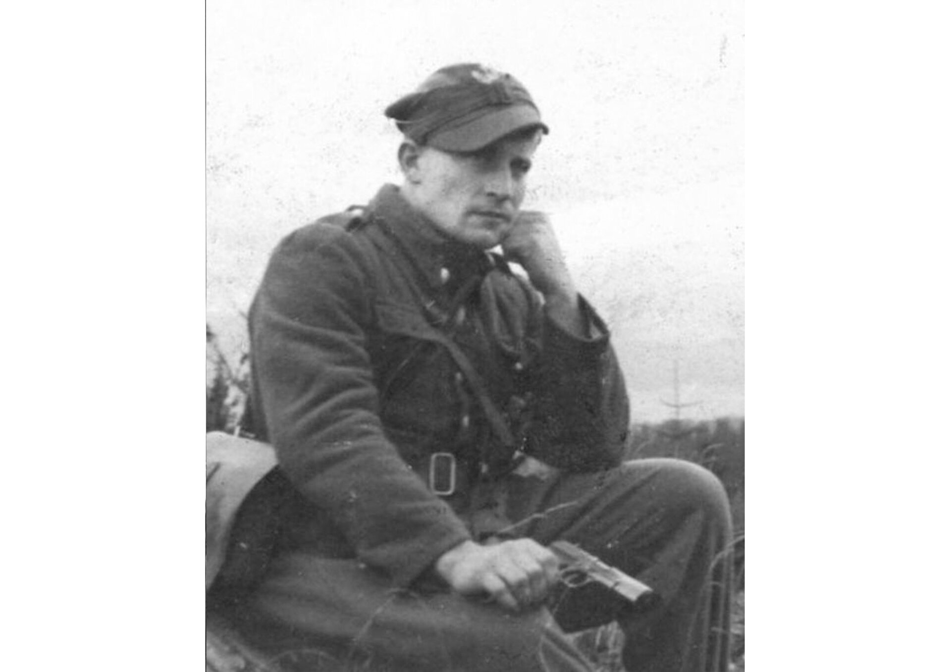 Fotografia portretowa Józefa Kurasi, pseudonim "Ogień". Mężczyzna w średnim wieku, ukazany w pozycji siedzącej. Trzyma w prawej ręce pistolet, lewa podpiera twarz. Ubrany w rogatywkę i wojskowy płaszcz.