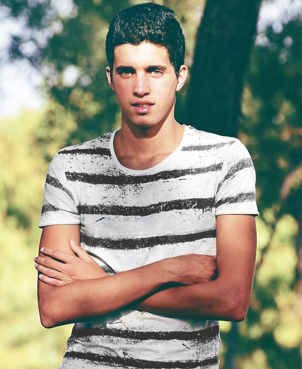 Zdjęcie przedstawia młodego mężczyznę ubranego w koszulkę, o ciemnej karnacji i czarnych włosach, trzymającego na brzuchu skrzyżowane ręce.