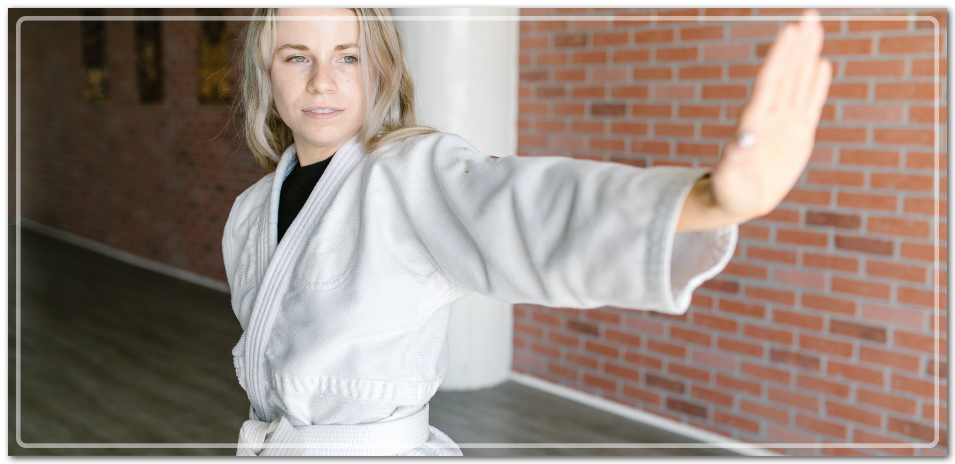 Ilustracja przedstawia kobietę z wyciągniętą przed siebie jedną ręką. Jest ona w białym kimonie wykorzystywanym w takich sportach jak na przykład karate. W pasie przewiązana jest białym pasem.