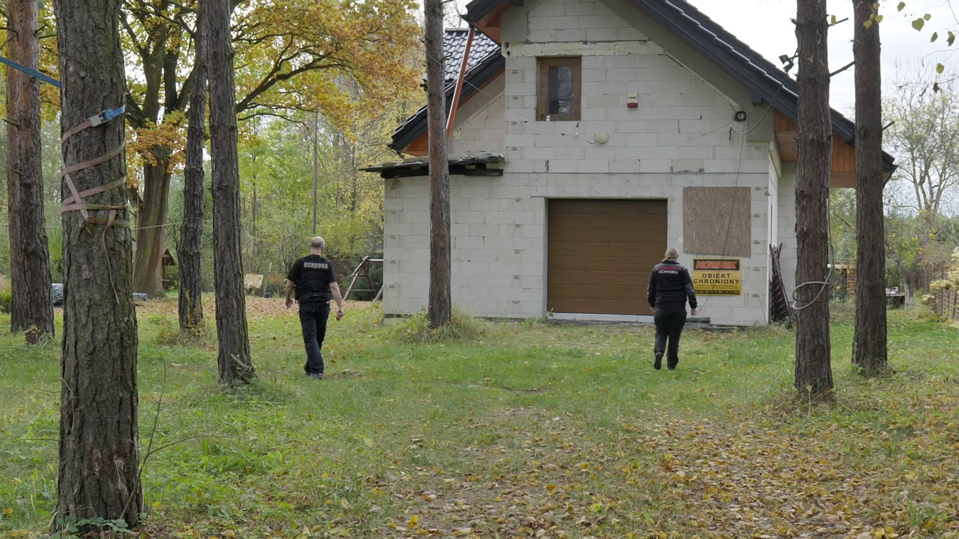 Zdjęcie pierwsze, podpisane ochrona obiektów, przedstawia dwóch ochroniarzy zbliżających się do domu w trakcie budowy. Dom znajduje się w lesie.