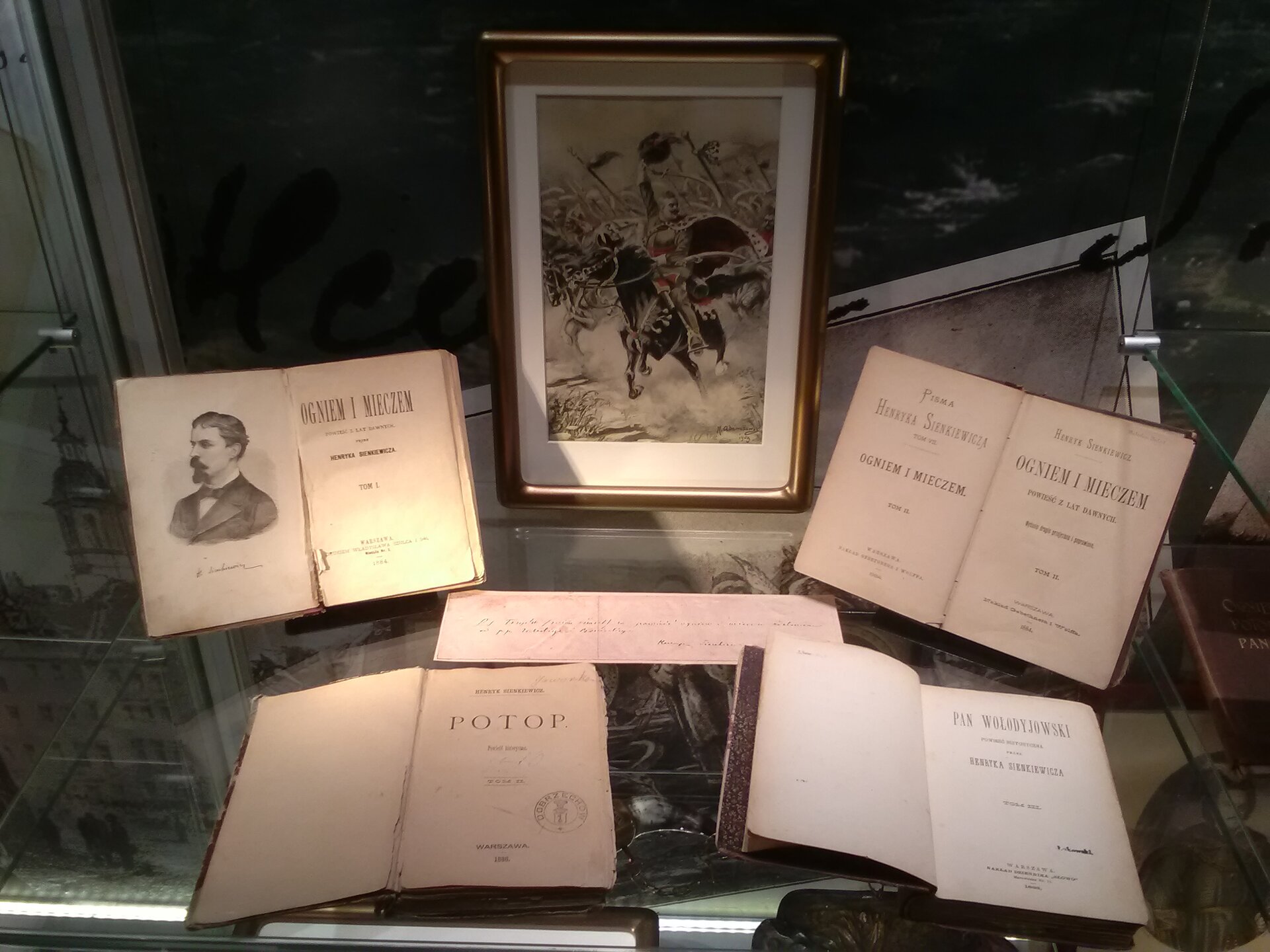 Zdjęcie przedstawia gablotę, w której znajduje się wystawa książek Henryka Sienkiewicza: "Ogniem i mieczem", "Potop" oraz "Pan Wołodyjowski". W gablocie stoi również oprawione w ramkę zdjęcie jeźdźcy na koniu.