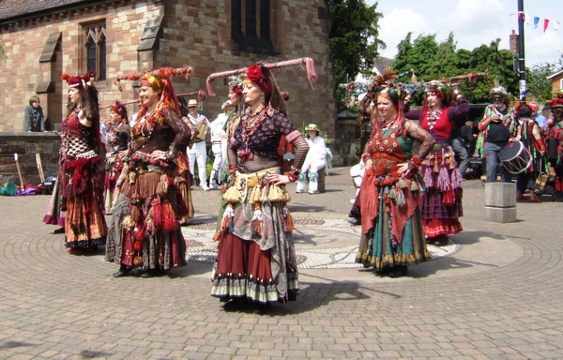 Fotografia przedstawiająca tańczące Romki ubrane w stroje etniczne. Kobiety mają na głowie czepce ozdobione kwiatami. Obcisłe bluzki i wzorzyste kamizelki oraz spódnice wykonane z warstwowego materiału bogato zdobione frendzlami. Kobiety tańczą w rzędach trzymając ręce pod bokami. Kobiety wykonują taniec na środku dużego placu, po lewej stronie stoi budynek z cegły.