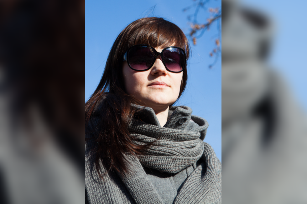 Fotografia przedstawiająca młodą dziewczynę w ciemnych okularach przeciwsłonecznych. Dziewczyna ma długie włosy z grzywką, ubrana jest w płaszcz ze stójką oraz grubo pleciony szary szalik zawiązany na płaszczu. W prawym górnym rogu ilustracji widać fragment drzewa. W tle bezchmurne niebo.