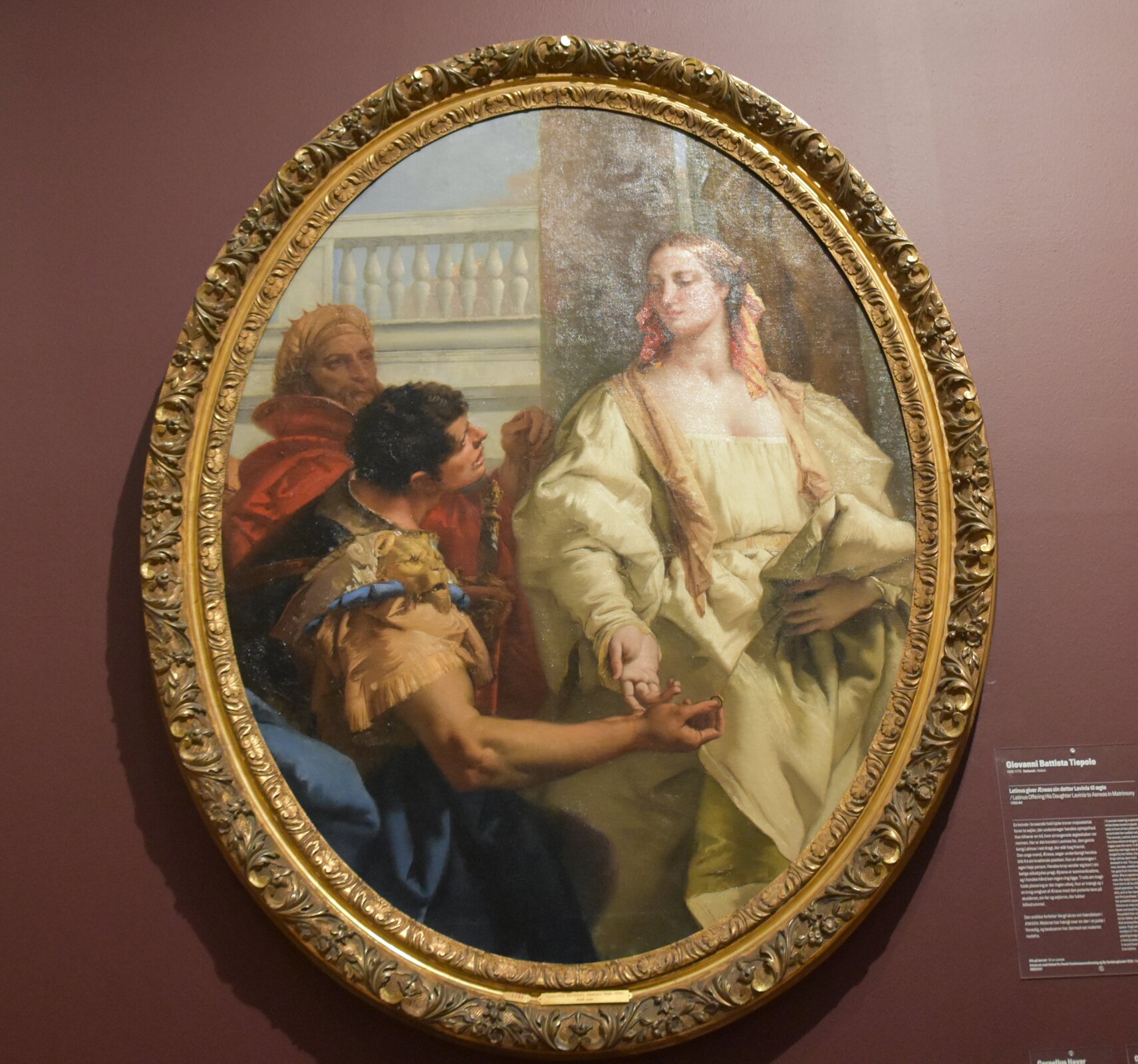 Ilustracja przedstawia obraz pt. „Latynus oferujący córkę Lawinię Eneaszowi za żonę”, którego autorem jest Giovanni Battista Tiepolo. Obraz ma kształt elipsy i jest w złotej, ozdobnej ramie. Po prawej stronie znajduje się kobieta o jasnych włosach. Ubrana jest w długą, kremową suknię z długim rękawem. W lewej dłoni trzyma dół sukni, a prawą dotyka dłoni mężczyzny. Mężczyzna ma niebieski strój z ozdobnymi, beżowymi rękawami. Ma krótkie, ciemne włosy. Za nim stoi starszy mężczyzna z żółtym nakryciem głowy i czerwonym strojem. W tle jest brązowa ściana i biała balustrada. Obraz znajduje się na śliwkowym tle.