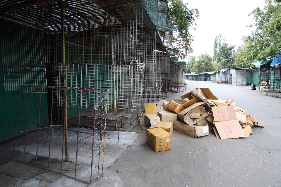 Zdjęcie przedstawia opustoszałe blaszane boksy handlowe stojące wzdłuż asfaltowej aleii. Są porozdzielane metalową siatką. Na alejce piętrzy się stos kartonowych pudeł. Są puste. 