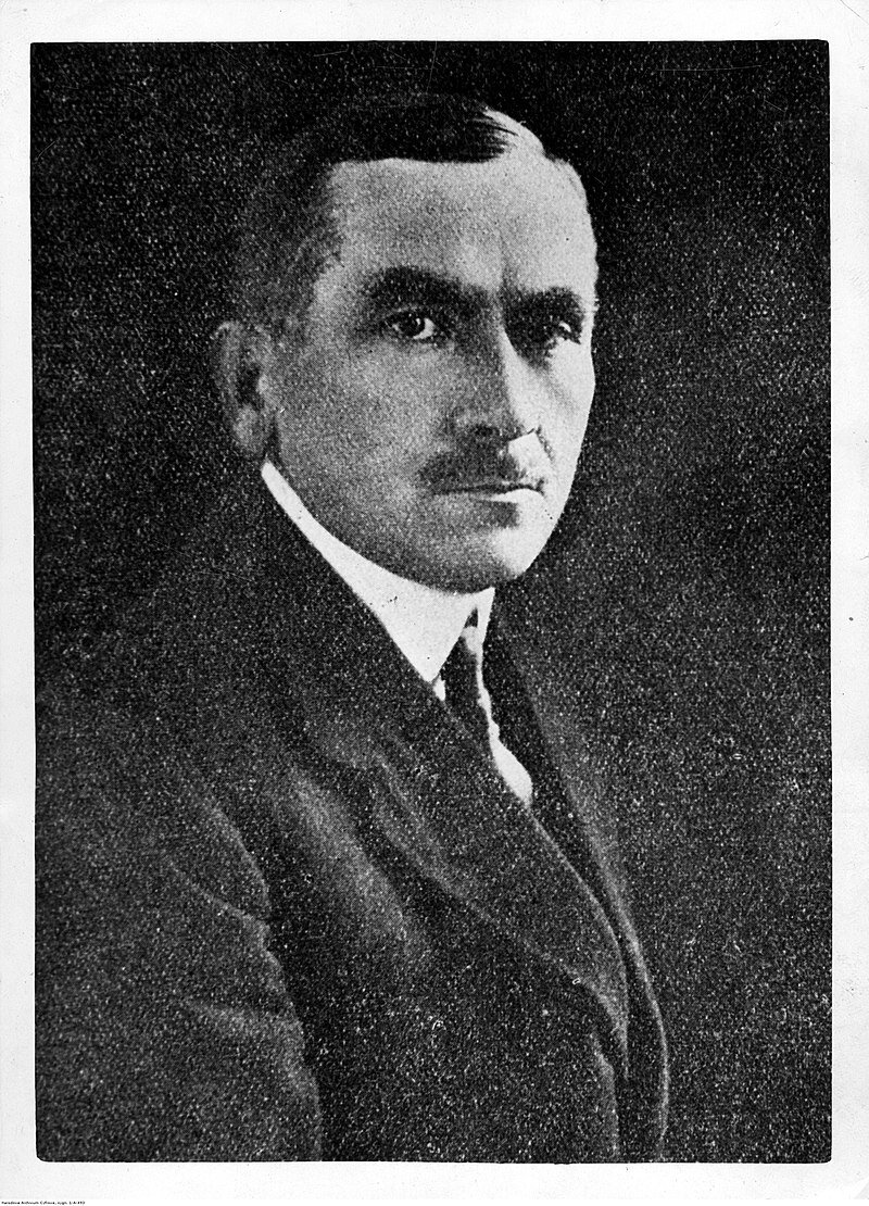 Fotografia przedstawia mężczyznę w średnim wieku. Widzimy głównie jego prawy profil. Mężczyzna ma ciemne, krótkie włosy zaczesane na bok, szczupłą twarz, wyraźnie zarysowane łuki brwiowe i ciemny, krótki wąsik. Ubrany jest w marynarkę, białą koszulę i krawat.  