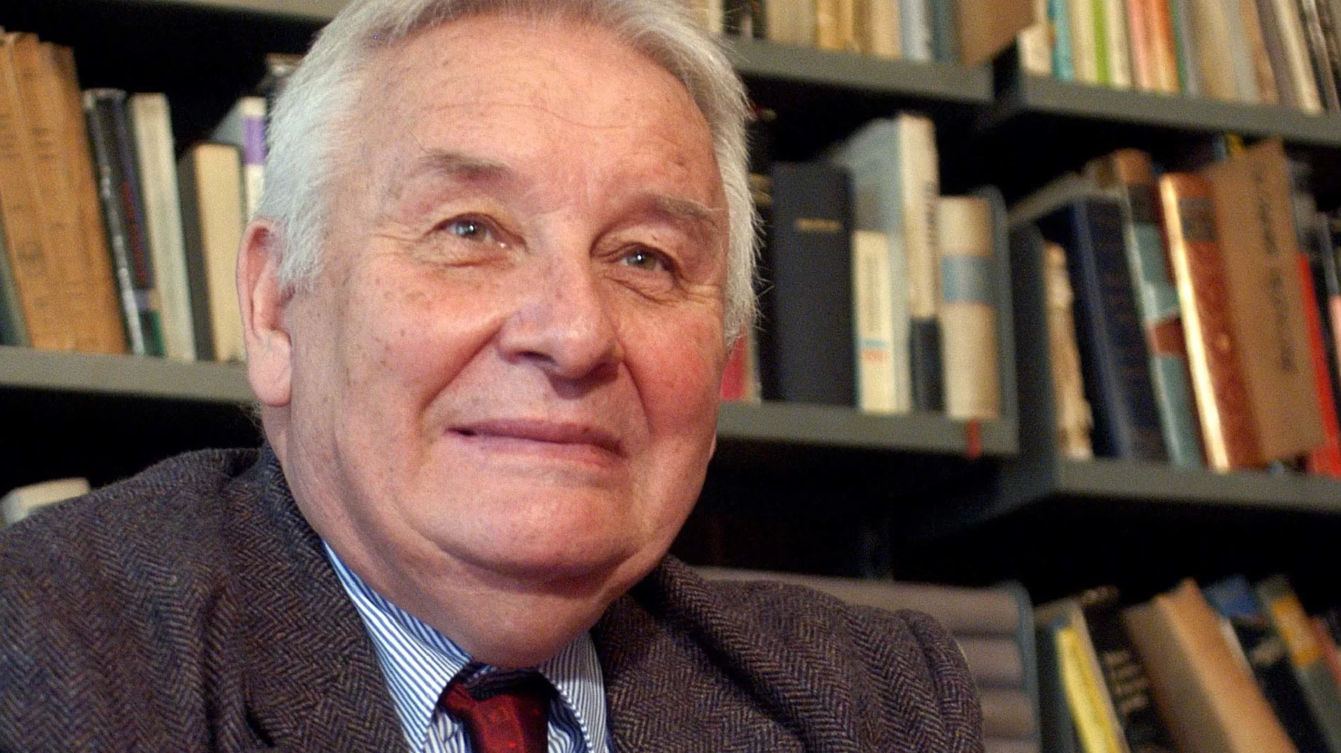 Ilustracja przedstawia fotografię uśmiechniętego Henryka Mikołaja Góreckiego. Mężczyzna ukazany jest na tle biblioteczki z książkami. 