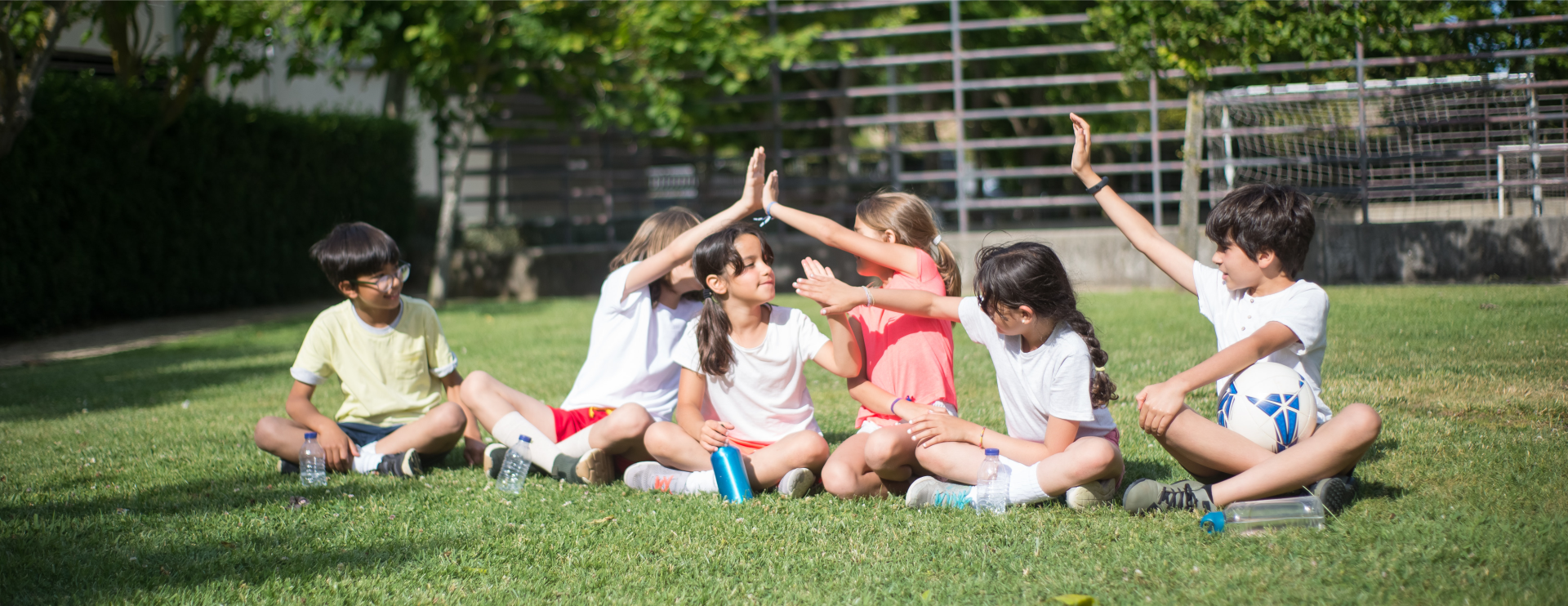 Ilustracja przedstawia gromadę zadowolonych dzieci, które siedzą na trawie. Przybijają ze sobą piątki.