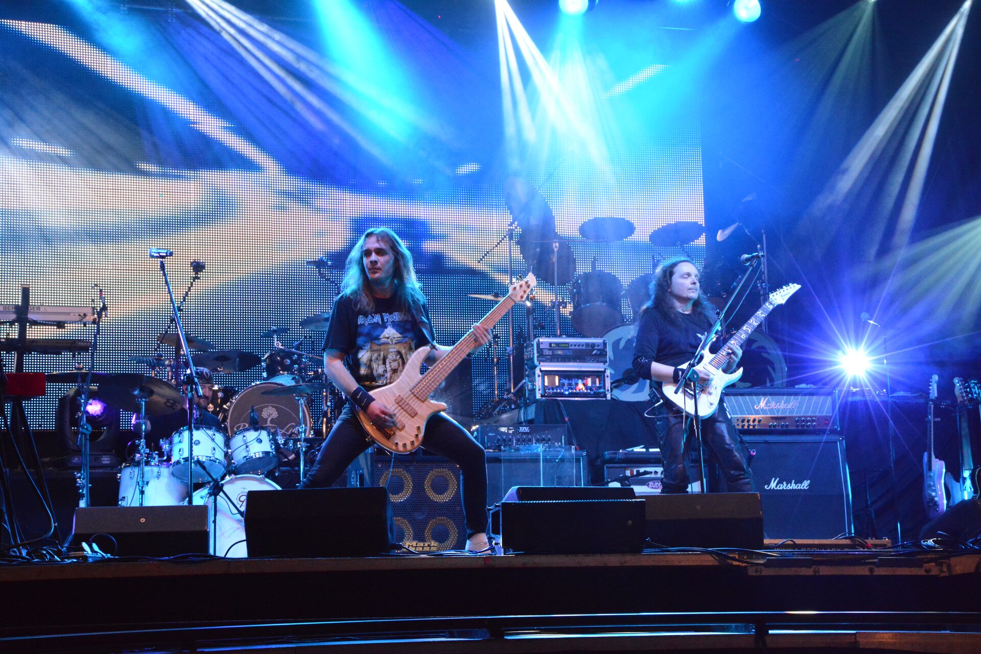 Ilustracja przedstawia koncert rockowy. Widocznych jest dwóch mężczyzn, grających na gitarach. W tle widoczna jest perkusja oraz głośniki.