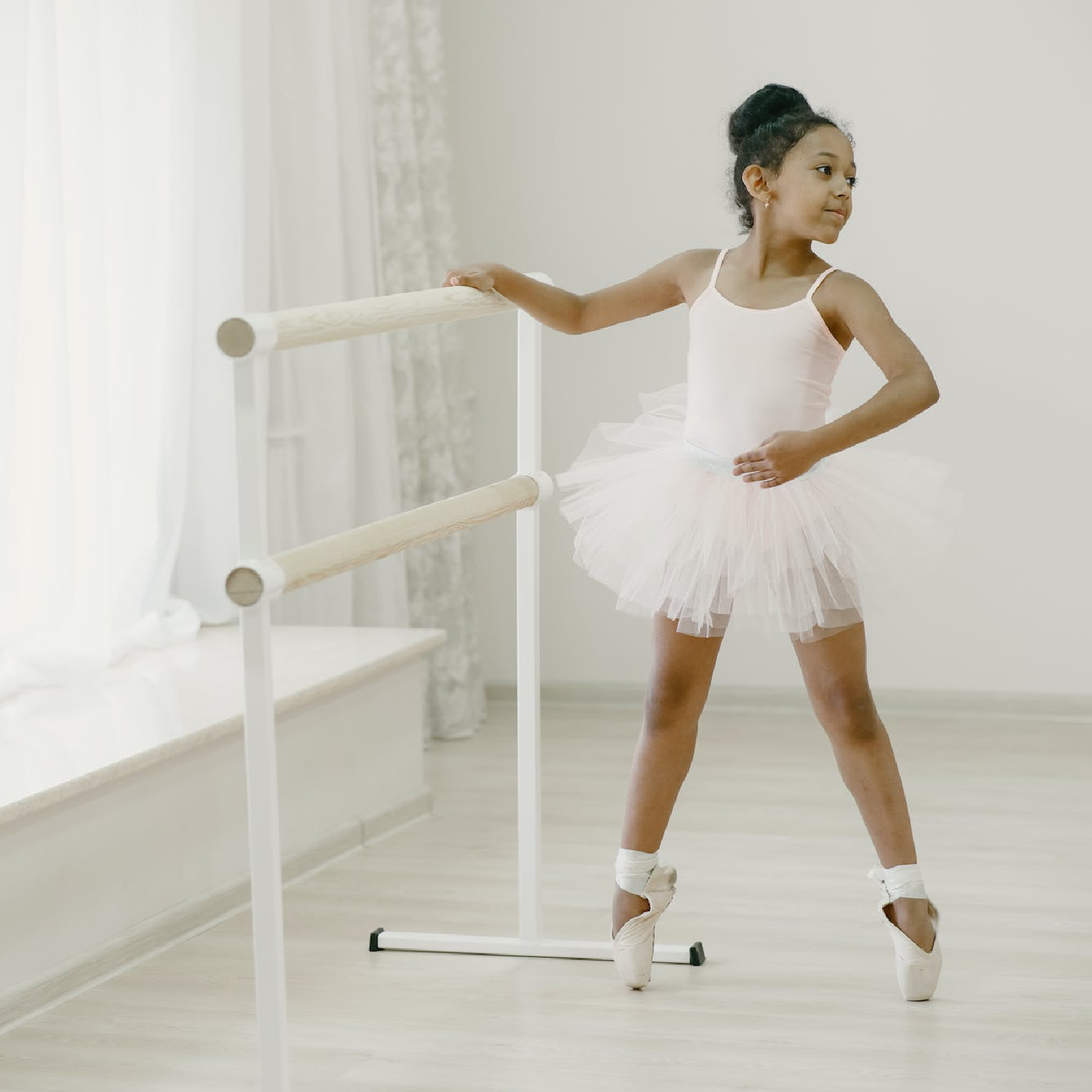 Ilustracja przedstawia małą dziewczynkę w wieku wczesnoszkolnym, w stroju baletnicy. Dziewczynka stoi na palcach stóp, opierając się o poręcz.