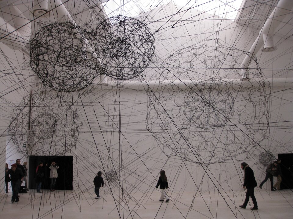 Zdjęcie ukazuje wnętrze wysokiego pomieszczenia, w którym znajduje się kilka osób w ciemnych ubraniach. Postacie są w jasnym, wysokim pomieszczeniu o jasnej podłodze. Nad ludźmi znajdują się 4 duże, ażurowe kule w kolorze czarnym. Niżej są 2 o mniejszej średnicy. Dodatkowo w przestrzeni pomieszczenia umieszczona została konstrukcja zbudowana z różnego rodzaju lin przybierająca kształt sieci. Liny biegną wzdłuż i wszerz pomieszczenia, kotwy mocujące konstrukcję widoczne są także na suficie i posadzce.