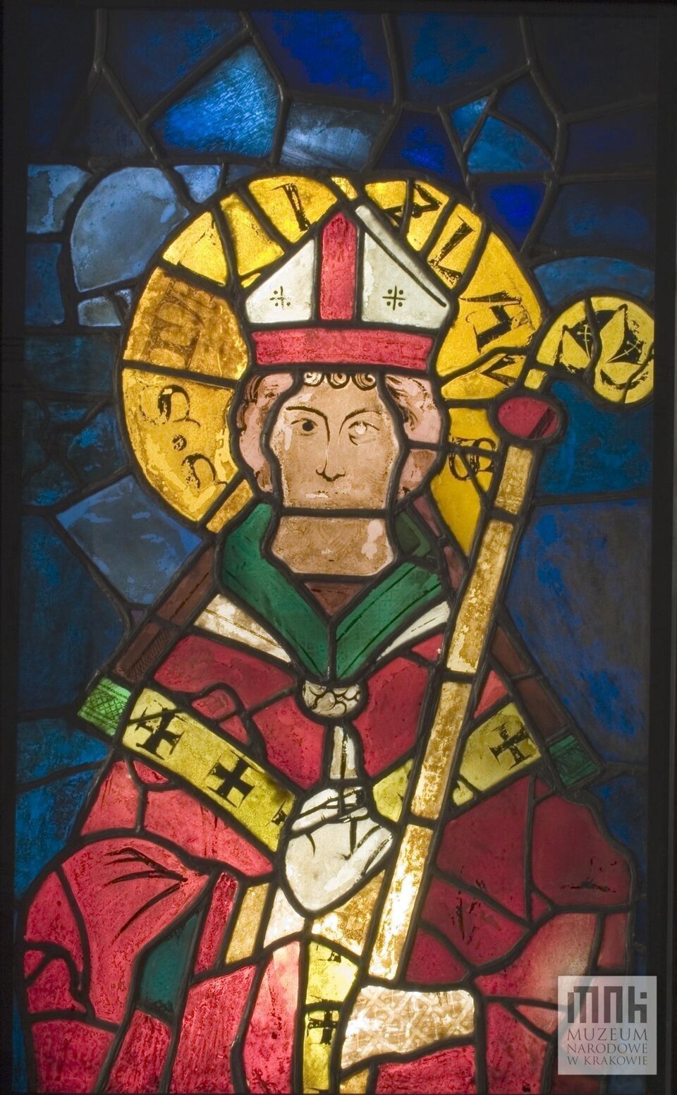 Ilustracja przedstawia witraż ze św. Stanisławem z kościoła Dominikanów z XIII w. Ukazuje świętego frontalnie. ubrany jest w strój biskupi, jego głowę otacza nimb. W ręku trzyma laskę biskupią. Tło witraża jest niebieskie.