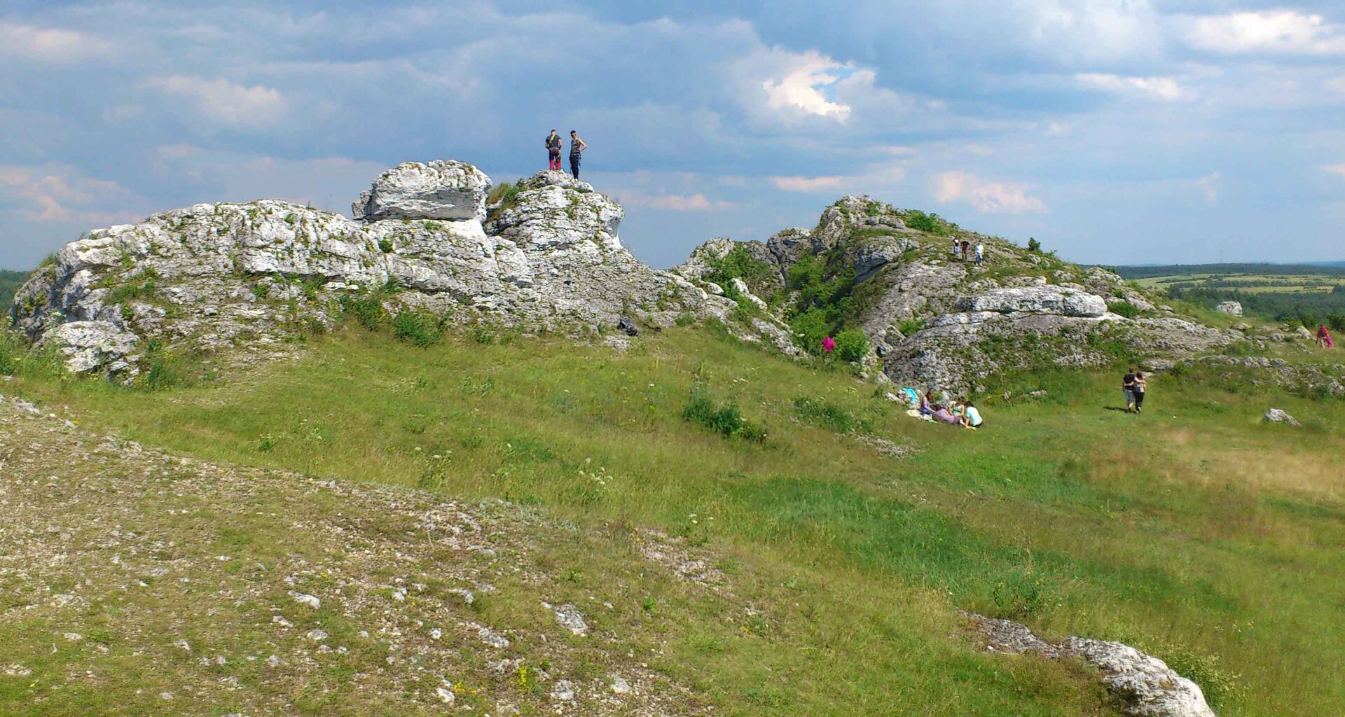 Zdjęcie pagórkowatego terenu pokrytego wapiennymi skałami. Na pierwszym planie dość łagodne zbocze porośnięte rzadką trawą lub porostami, przez które przebijają się gdzieniegdzie białe skały. Na drugim planie, w centralnej części zdjęcia, wystające na kilkanaście metrów białe spękane skały o nieregularnym kształcie, w niektórych miejscach pokryte drobną roślinnością, najczęściej trawą lub niewielkimi krzewami. Na skałach, a także przed nimi spacerują ludzie. W tle rozległy teren wyżynny, położony nieco niżej względem pierwszego planu.