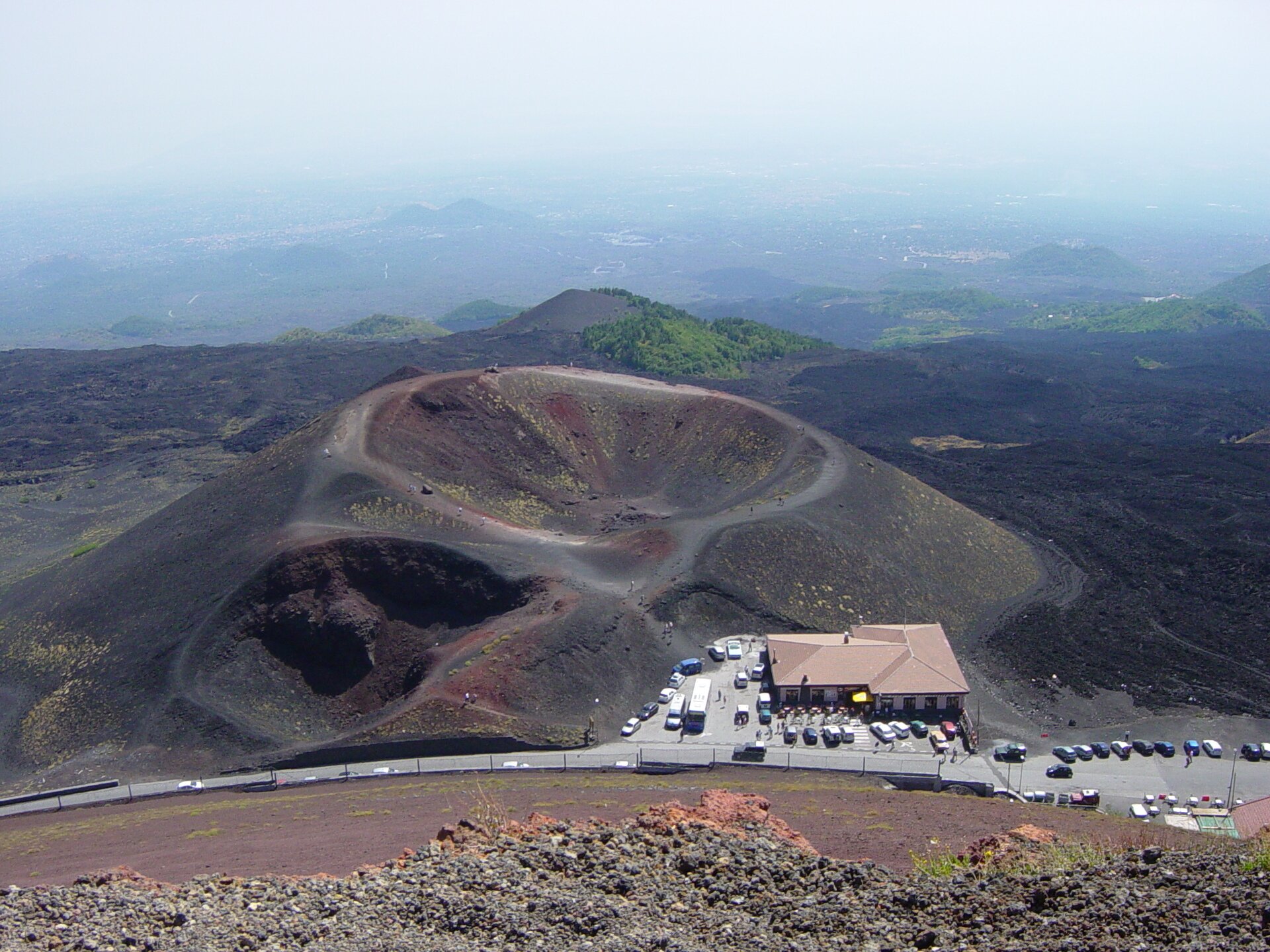 Na zdjęciu lotniczym znajduje się wulkan Etna. W centralnej części zdjęcia widoczny jest wklęsły krater. Jest to okrągłe wgłębienie. Zbocza wulkanu są zbudowane z czarnych skał wulkanicznych oraz odłamków zastygłej lawy. Na drugim planie widoczny jest surowy, skalisty krajobraz wulkaniczny, gdzieniegdzie porośnięty zieloną roślinnością. Niedaleko krateru widać parking dla samochodów i budynek.