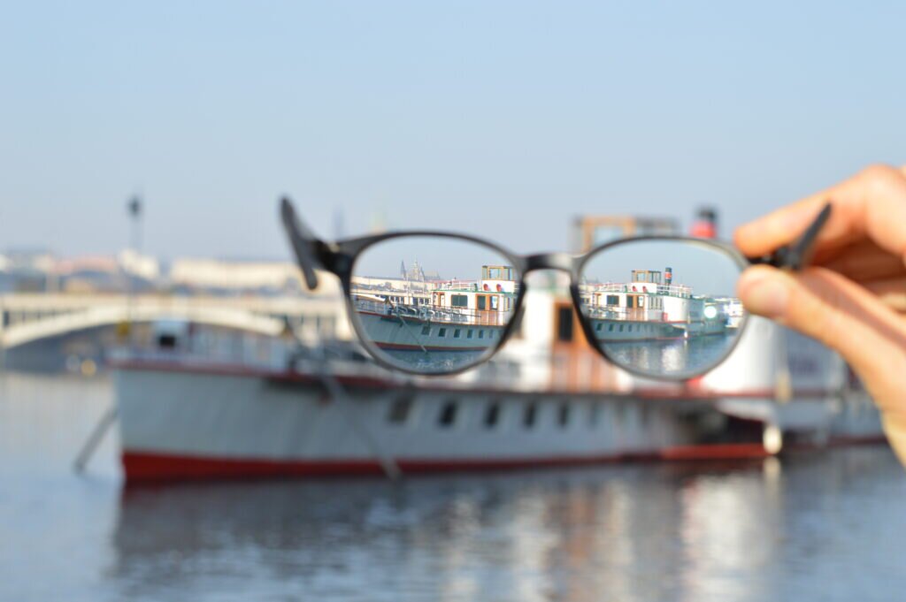 Zdjęcie przedstawia statek na wodzie. Obraz jest rozmazany. Na pierwszym planie znajdują się okulary korygujące trzymane przez osobę stojącą za aparatem. Obraz widziany przez szkła okularów jest prawidłowy.