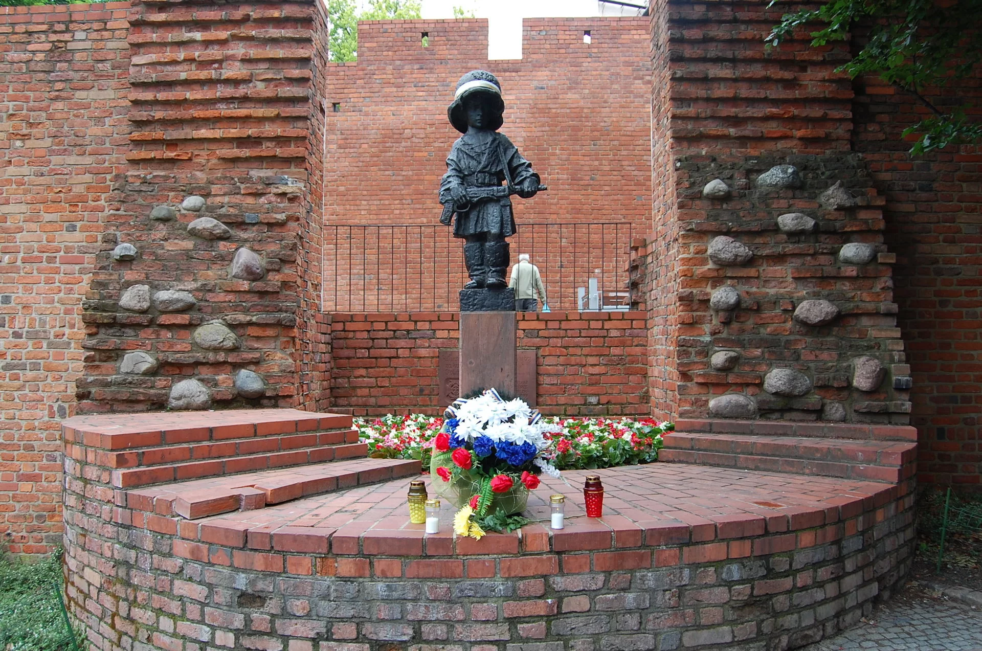 Kliknij, aby powiększyć Zdjęcie przedstawia pomnik dziecka ubranego w za duży płaszcz spięty w pasie paskiem, za duży hełm z otokiem w barwach narodowych (biało‑czerwonych) i za duże wysokie buty. Trzyma w rękach karabin przewieszony na pasku przez ramię. Pomnik ustawiony jest na cokole, który stoi na ceglanym półkolistym murku. Po obydwu stronach oraz za pomnikiem widać ceglane ściany.