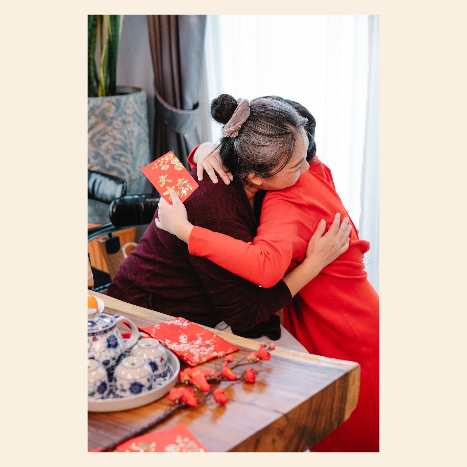 Ilustracja przedstawia babcię z fryzurą w kok przytulającą swoją wnuczkę, która trzyma w dłoni kartkę podarunkową. Za kobietami widać okno oraz kwiatek doniczkowy. Przed kobietami znajduje się stół z zestawem filiżanek i kwiatkami.