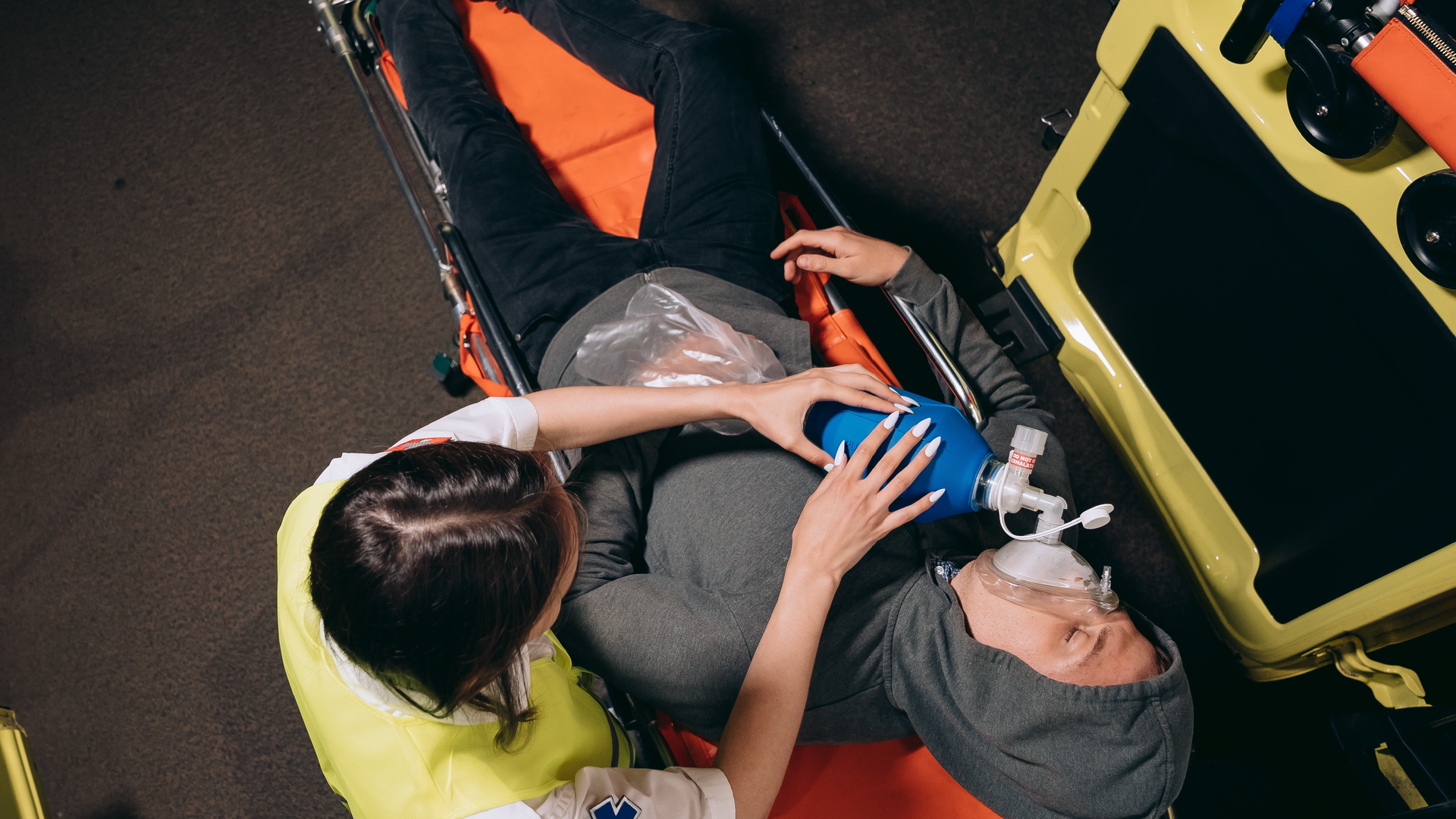 Zdjęcie przedstawia kobietę udzielającą pierwszej pomocy mężczyźnie. Mężczyzna leży na noszach w bluzie z kapturem założonym na głowie. Na twarzy mężczyzna ma maskę tlenową. Kobieta nad nim trzyma aparat z gruszką i maską do sztucznego oddychania.
