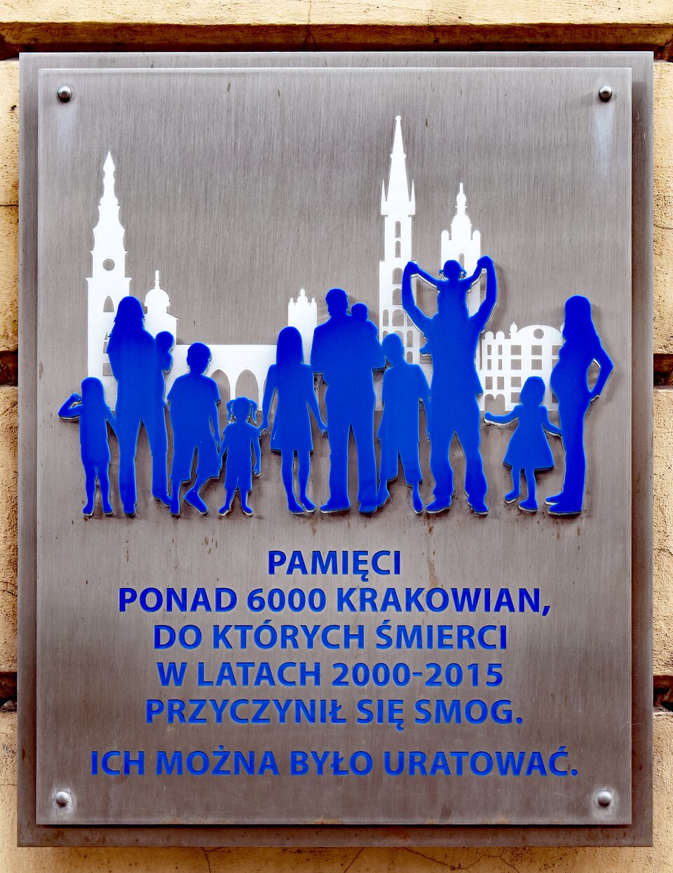 Na ilustracji ukazano metalową tablicę, na której znajdują się niebieskie sylwetki ludzi, a za nimi białe budynki. Pod spodem znajduje się niebieski napis: Pamięci ponad sześciu tysięcy krakowian, do których śmierci w latach dwa tysiące dwa tysiące piętnaście przyczynił się smog. Ich można było uratować.