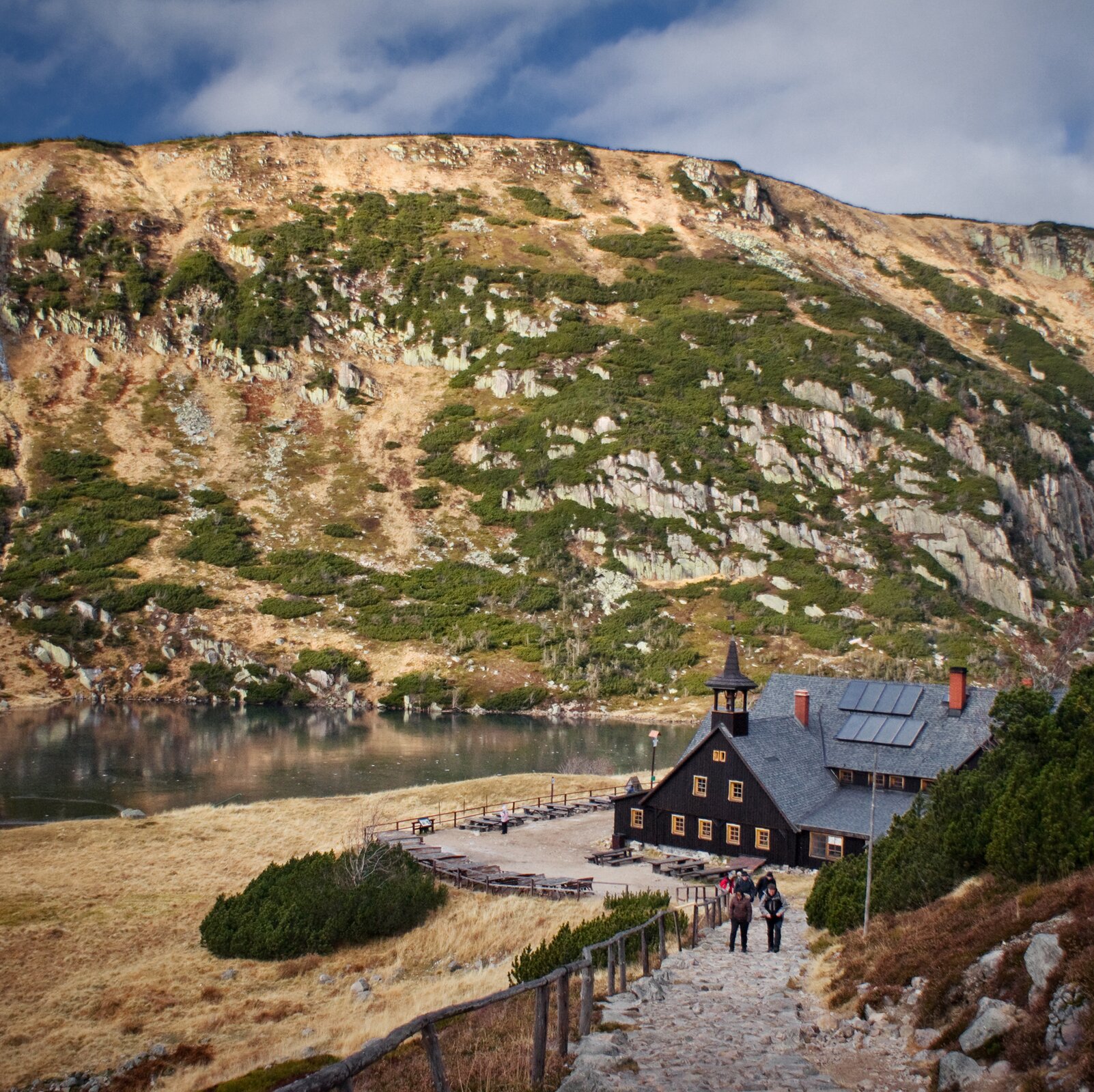 Fotografia. Na pierwszym planie droga wyłożona kamieniami prowadząca wprost do schroniska położonego tuż przy niewielkim jeziorze. Zbiornik wodny znajduje się u podnóża wzniesienia, które jest na drugim planie. Wysokie wzniesienie pokryte gdzieniegdzie krzewami, przebijają się przez nie jasne skały. Schronisko posiada spadzisty dach, ma czarne ściany i jasnobrązowe okna. Przed budynkiem plac, na którym stoją stoły z ławkami.