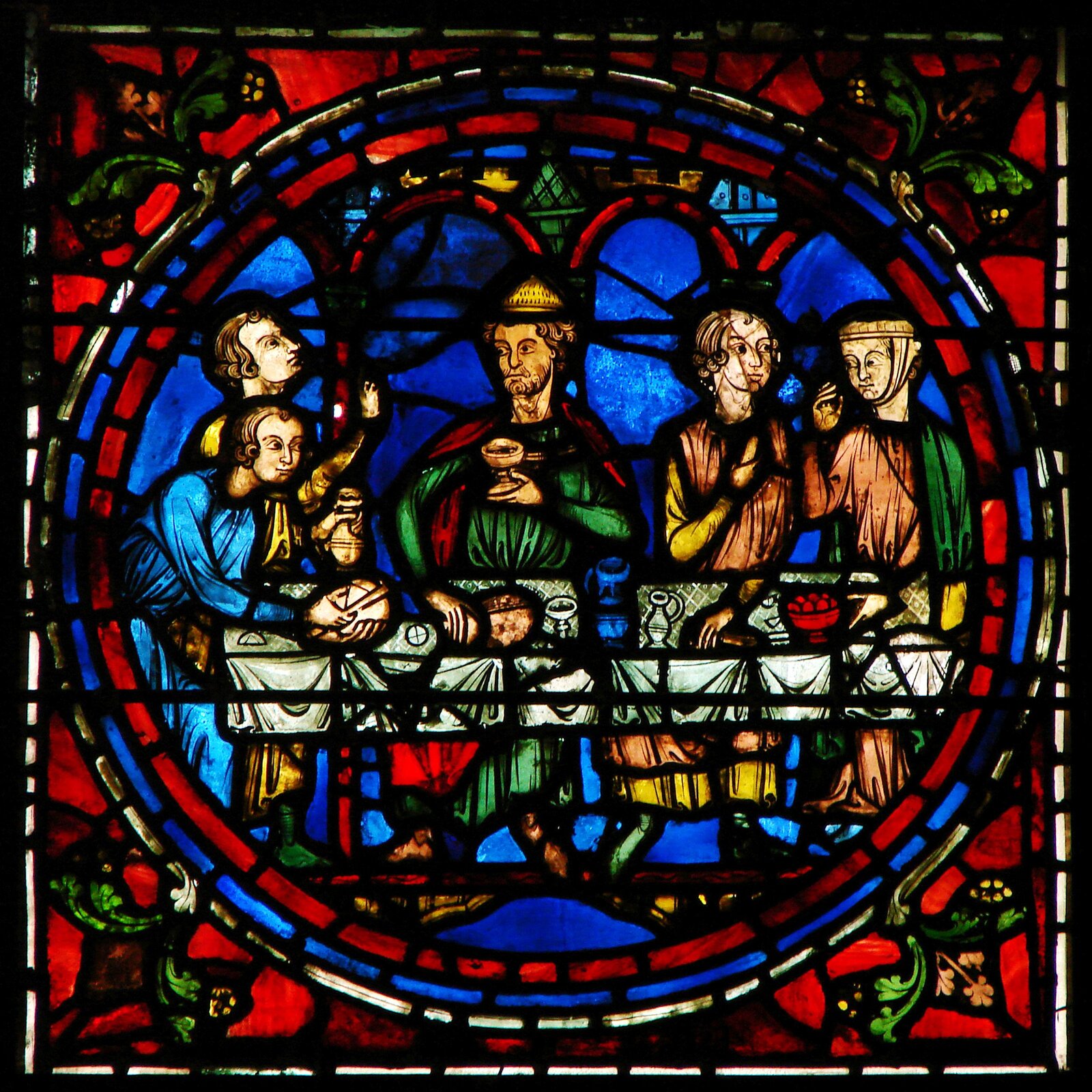 Zdjęcie przedstawia witraż z Katedry w Chartres z XIII wieku. Ukazuje Chrystusa i Apostołów, siedzących za stołem podczas Ostatniej Wieczerzy, przedstawionym w kole. Jezus trzyma kielich z winem. Po obu stronach znajdują się po dwie postacie. Tło jest niebieskie, a przestrzeń dookoła wypełniona jest czerwonymi szkiełkami. W narożnikach znajdują się motywy roślinne.