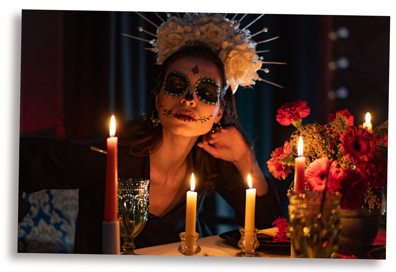 Ilustracja przedstawia kobietę z kwiatami na głowie i makijażem w stylu czaszki. Na pierwszym planie widoczne kwiaty i świece ustawione na stoliku.